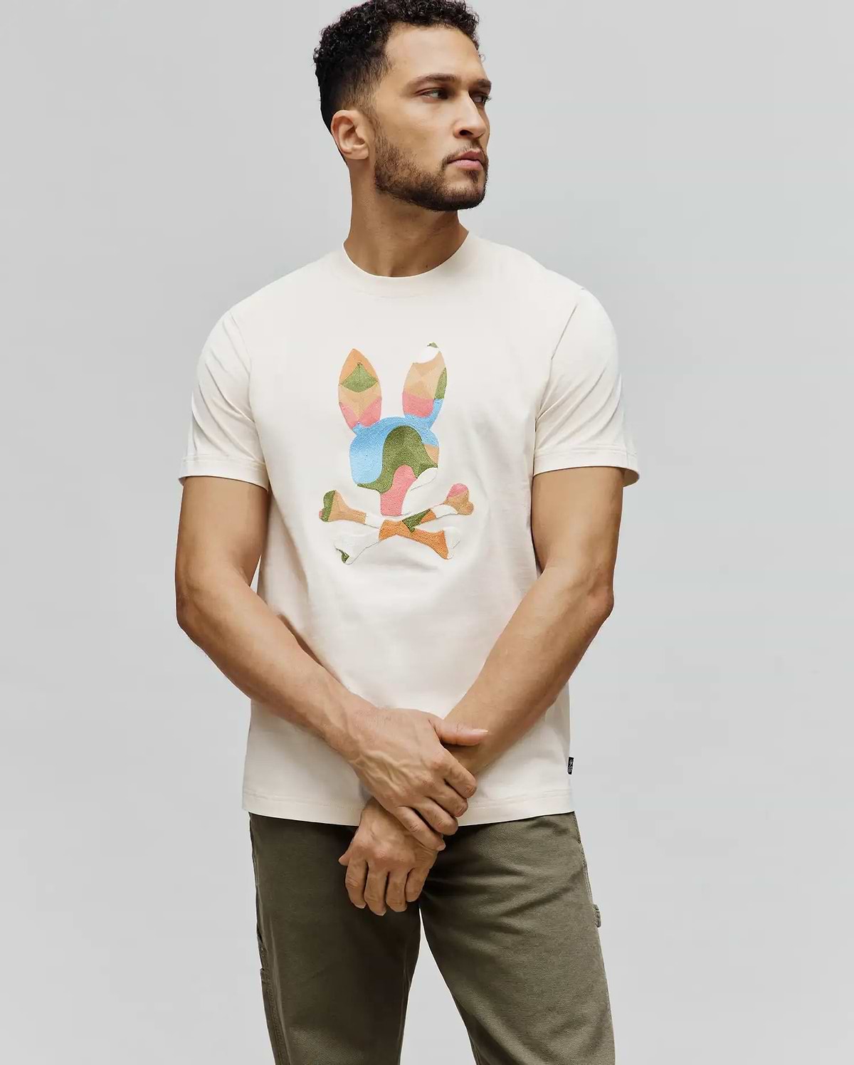 Cream Mens Ara Graphic Tee, classic fit jersey t-shirt featuring a large, geometric multi-color Psycho Bunny skull and crossbones graphic on the chest, with a small brand tag on the hem.