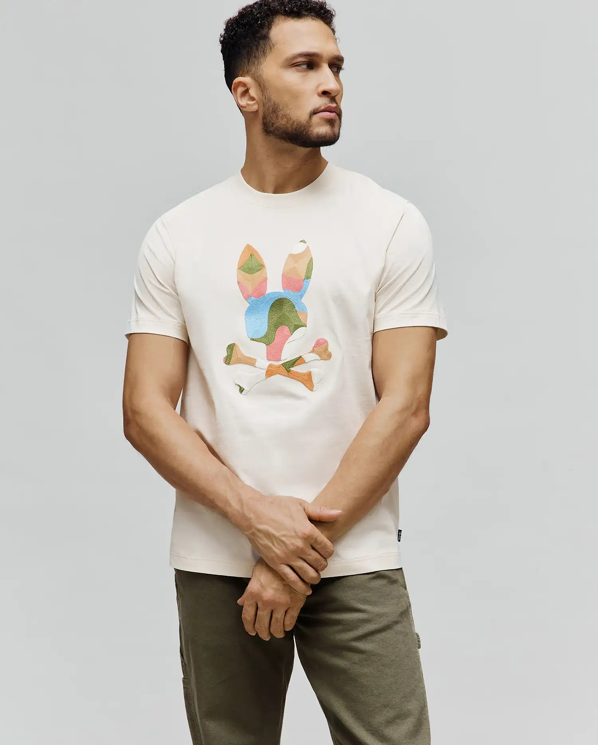Cream Mens Ara Graphic Tee, classic fit jersey t-shirt featuring a large, geometric multi-color Psycho Bunny skull and crossbones graphic on the chest, with a small brand tag on the hem.