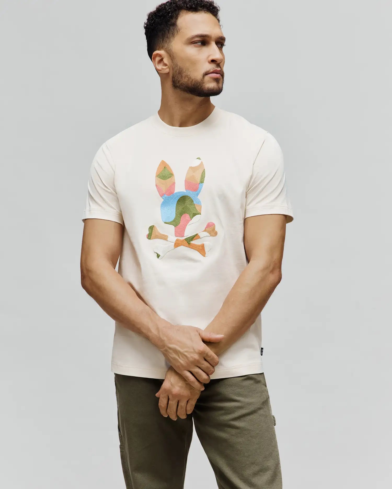 Cream Mens Ara Graphic Tee, classic fit jersey t-shirt featuring a large, geometric multi-color Psycho Bunny skull and crossbones graphic on the chest, with a small brand tag on the hem.