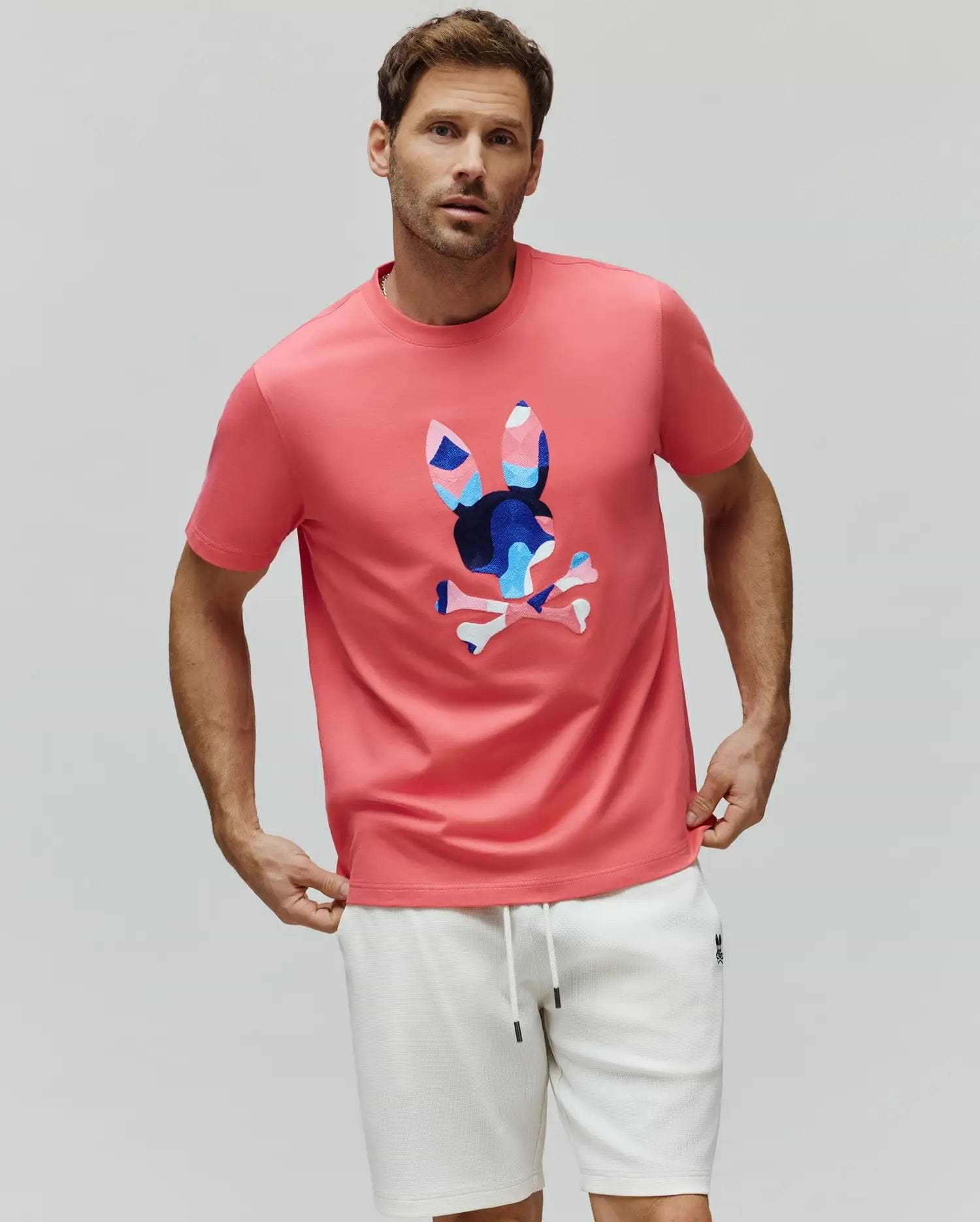 Mens Ara Graphic Tee, coral pink classic fit jersey knit t-shirt with a large, centered geometric Psycho Bunny skull logo in blue, light blue, and pink.