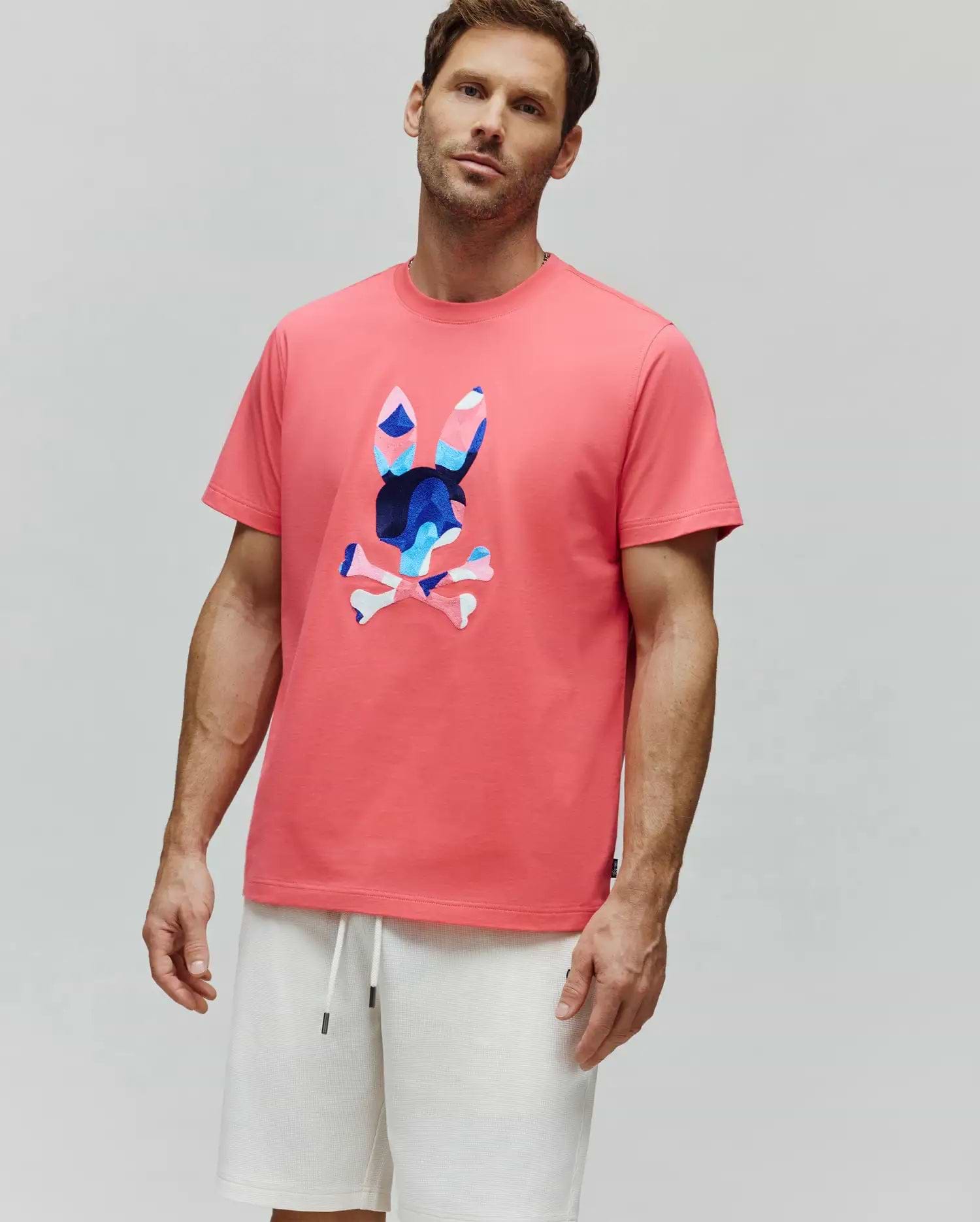 Coral pink Mens Ara Graphic Tee, classic fit jersey, with a large central Psycho Bunny skull logo featuring a vibrant blue, pink, and white camouflage pattern.