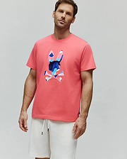 Coral pink Mens Ara Graphic Tee, classic fit jersey, with a large central Psycho Bunny skull logo featuring a vibrant blue, pink, and white camouflage pattern.