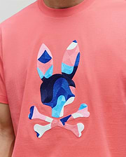 MENS ARA GRAPHIC TEE in coral jersey knit, featuring a large, central embroidered Psycho Bunny skull logo with geometric blue, pink, and white accents.