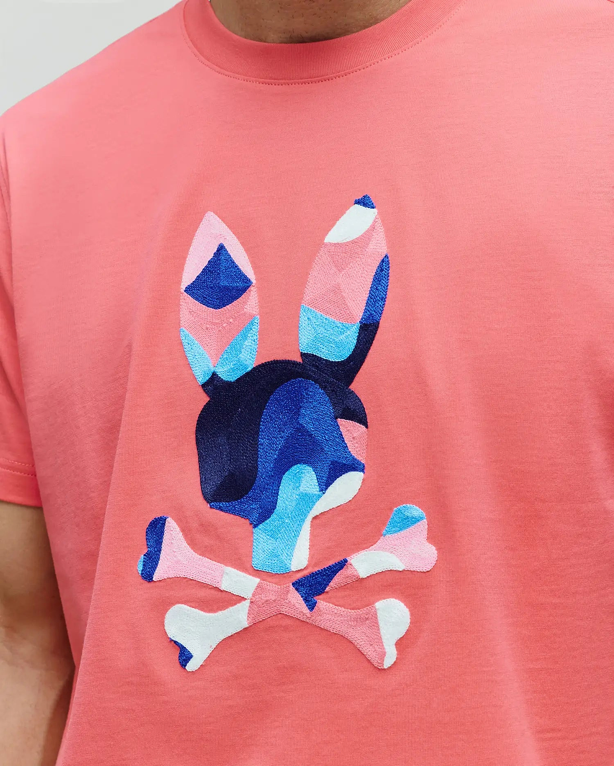MENS ARA GRAPHIC TEE in coral jersey knit, featuring a large, central embroidered Psycho Bunny skull logo with geometric blue, pink, and white accents.