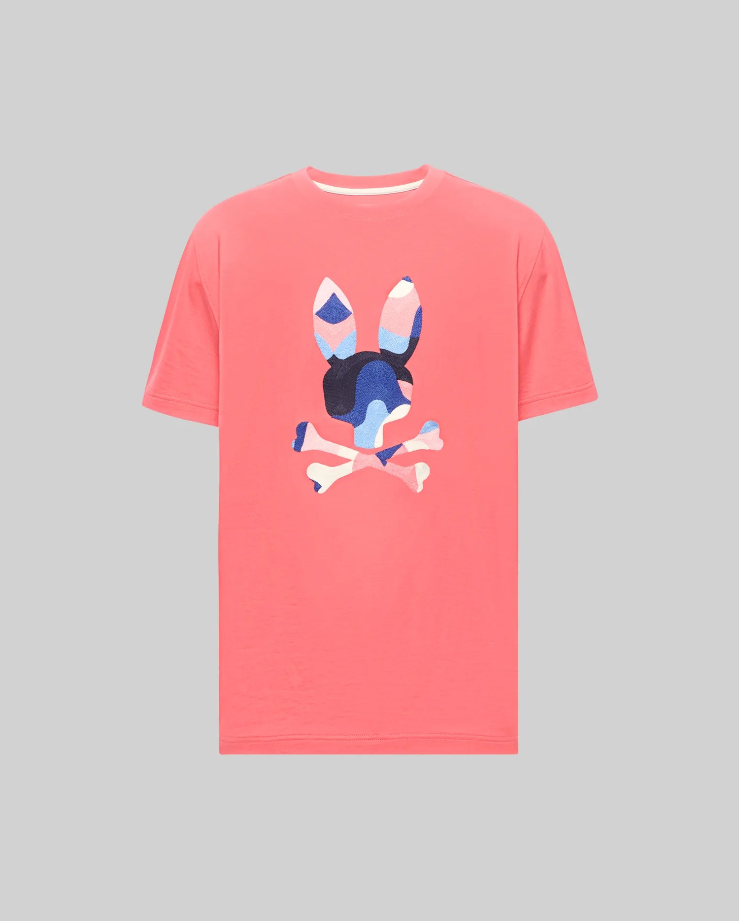 Mens Ara Graphic Tee in coral pink jersey, featuring a large central Psycho Bunny skull and crossbones logo with a vibrant blue, pink, and white abstract camouflage pattern, classic fit.