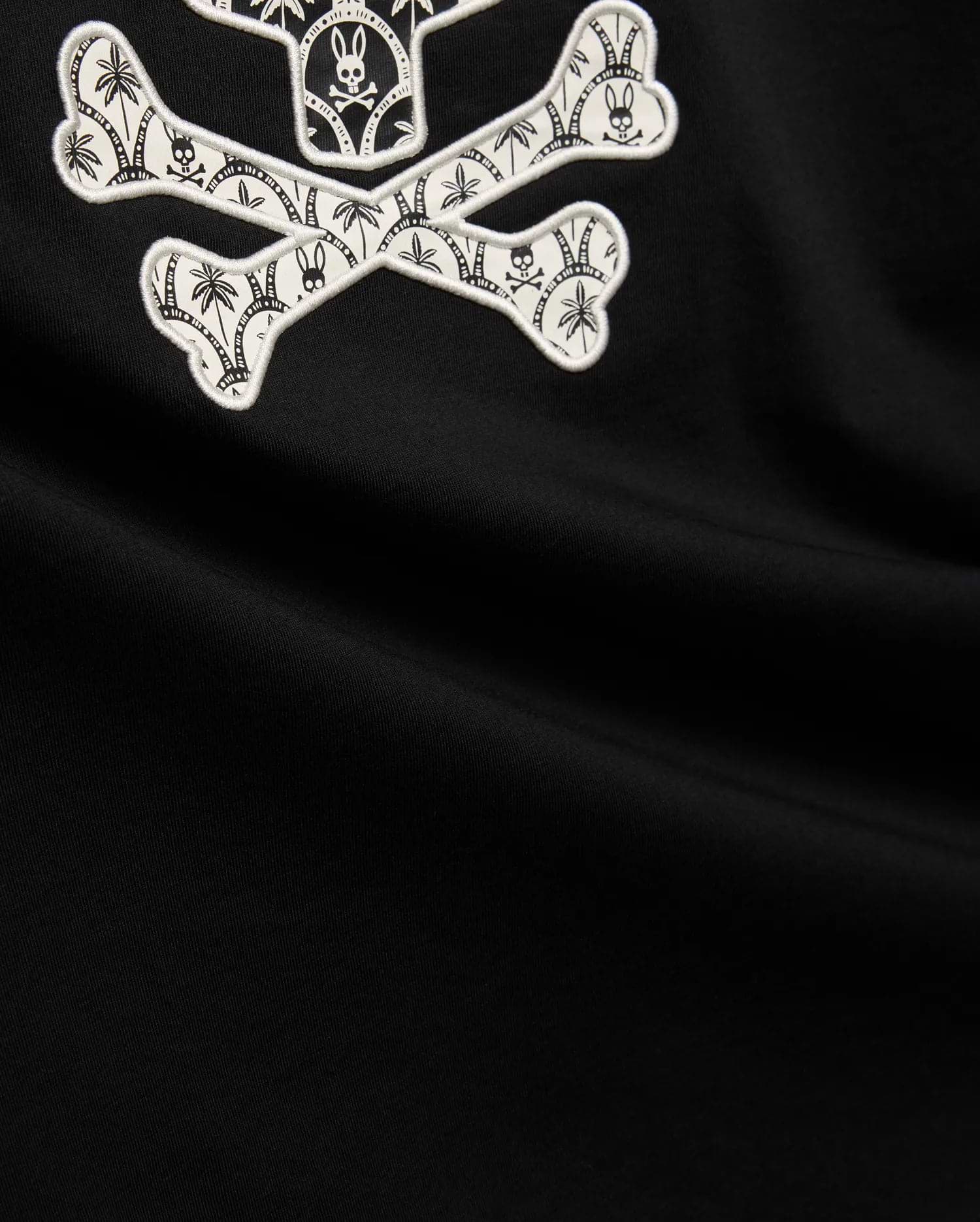 Mens Brad Graphic Tee, black smooth jersey with a large white embroidered Psycho Bunny skull and crossbones graphic, featuring tropical palm trees and mini bunny skulls within the bones.