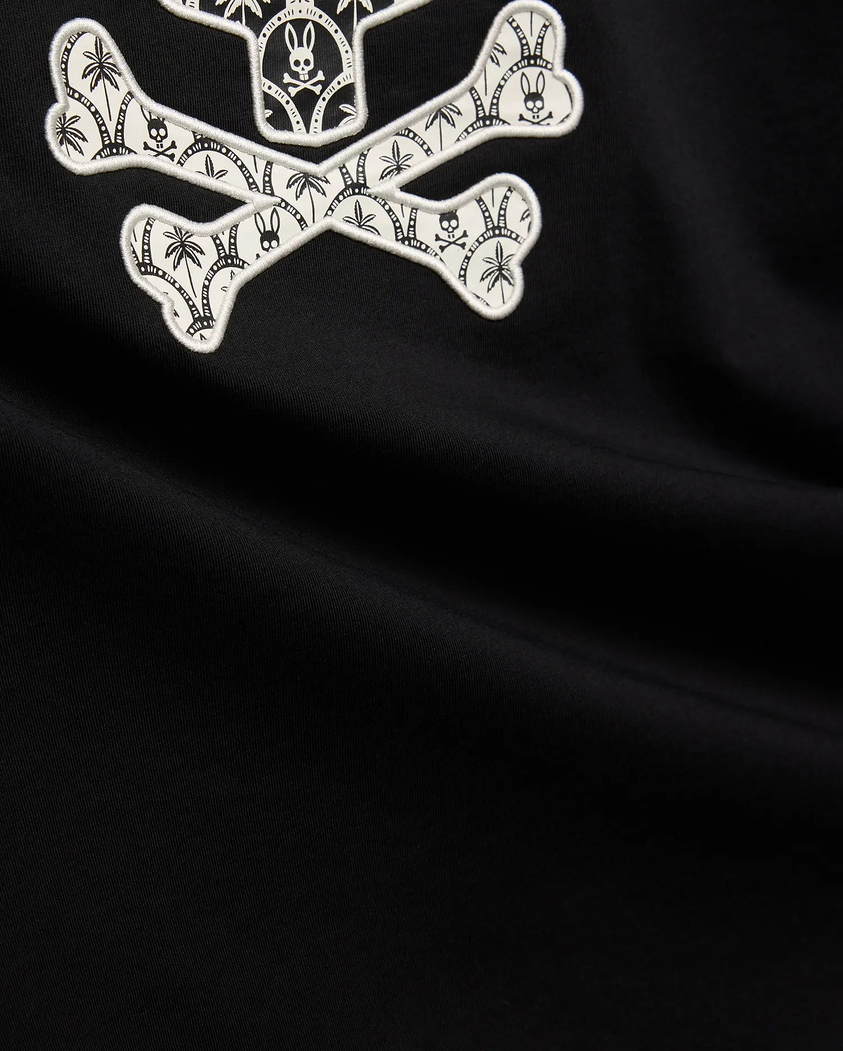 Mens Brad Graphic Tee, black smooth jersey with a large white embroidered Psycho Bunny skull and crossbones graphic, featuring tropical palm trees and mini bunny skulls within the bones.