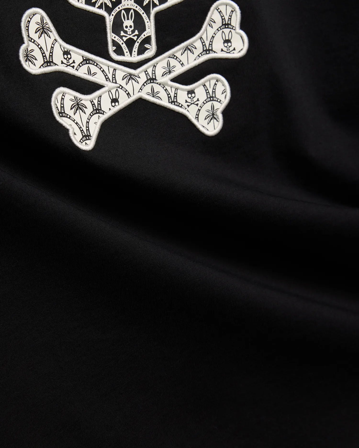 Mens Brad Graphic Tee, black smooth jersey with a large white embroidered Psycho Bunny skull and crossbones graphic, featuring tropical palm trees and mini bunny skulls within the bones.