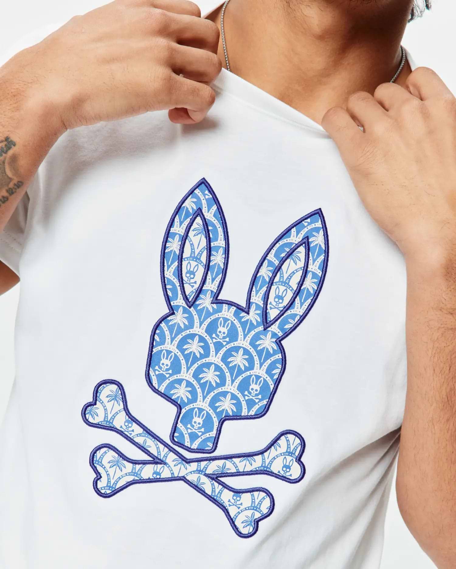 White Mens Brad Graphic Tee in soft jersey, featuring a large central Psycho Bunny skull logo with a blue and white palm tree and mini bunny skull pattern, outlined in dark blue embroidery.