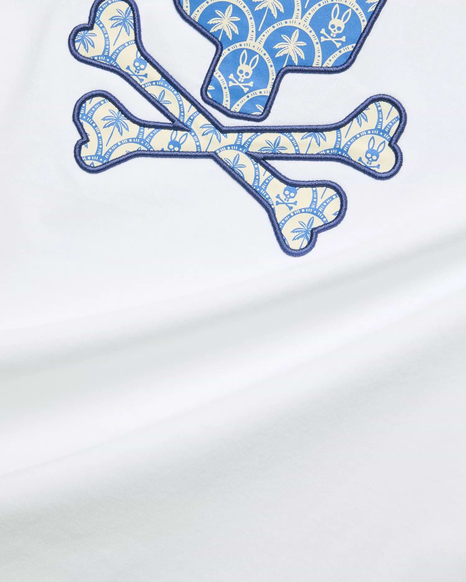 White Mens Brad Graphic Tee, classic fit, smooth jersey, featuring a large central Psycho Bunny skull and crossbones graphic with blue and cream palm tree and mini bunny skull pattern, outlined in dark blue embroidery.