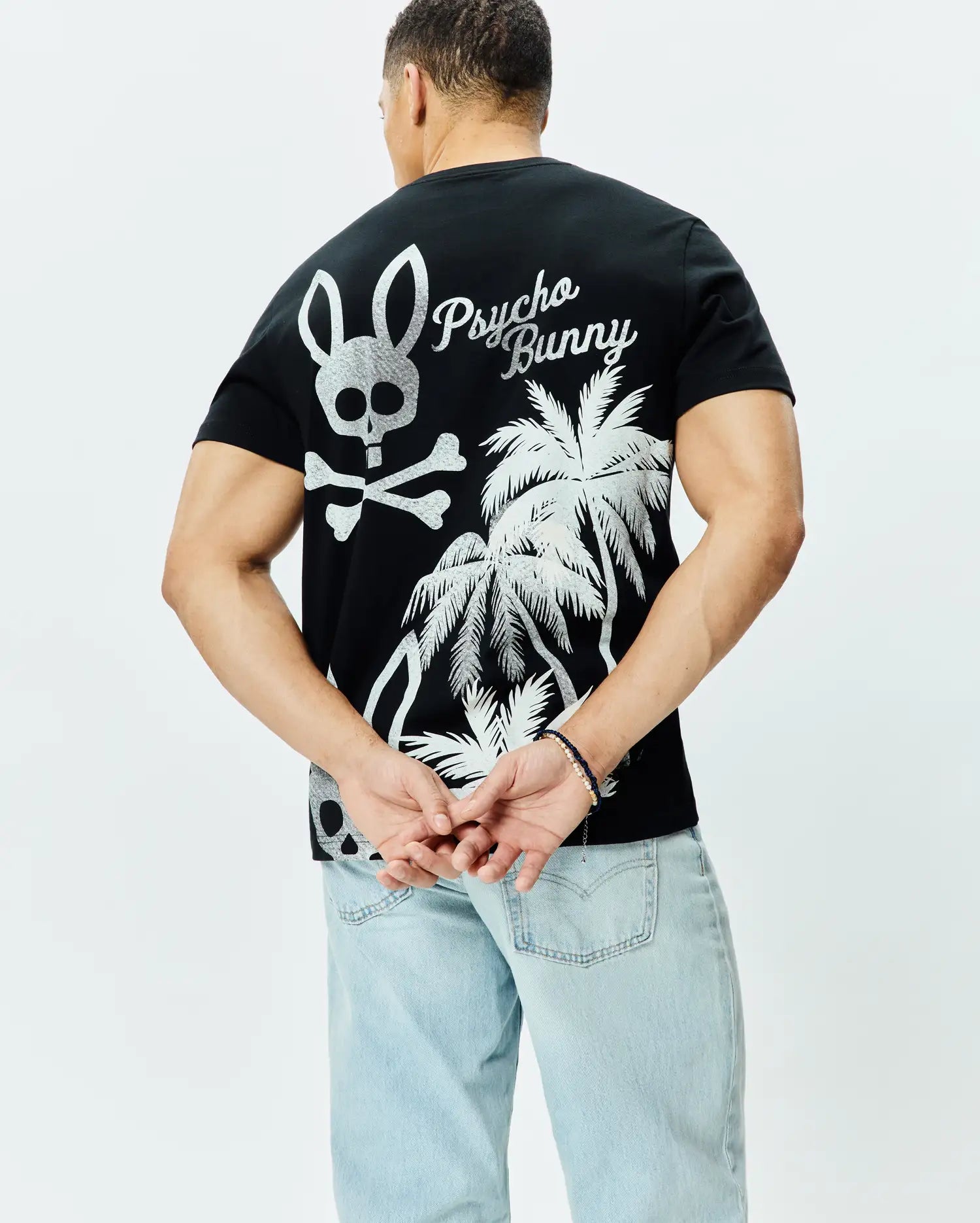 Mens Colin Back Graphic Tee, black jersey knit, classic fit, featuring a large silver Psycho Bunny skull and crossbones, script logo, and palm tree graphic on the back.