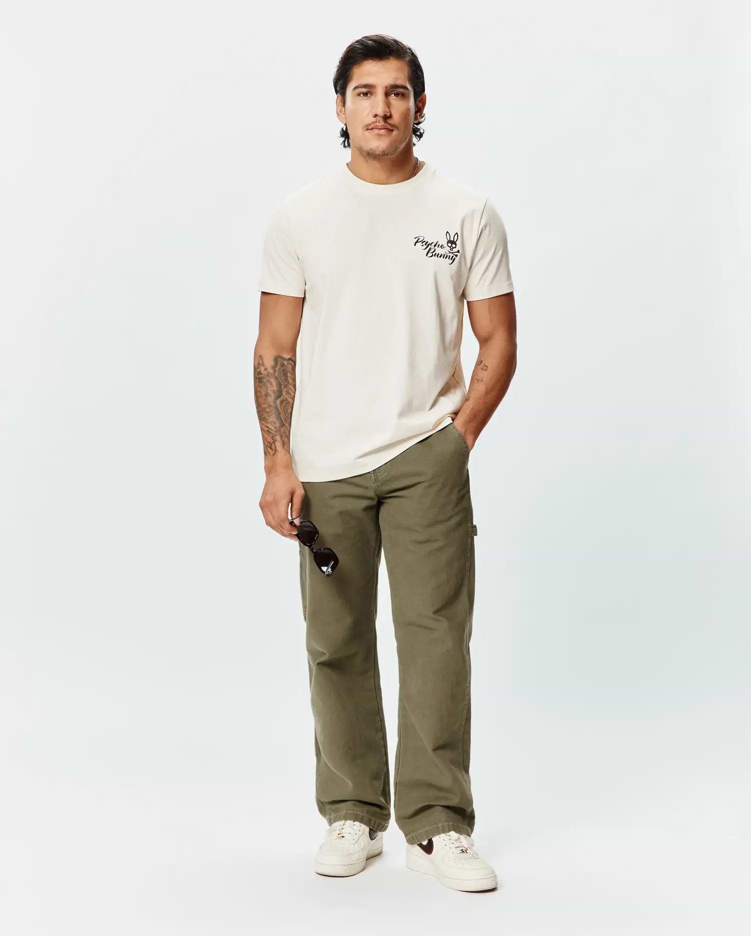 Mens Colin Back Graphic Tee, a classic fit ecru jersey t-shirt featuring a small, bold black Psycho Bunny skull logo and text printed on the left chest.