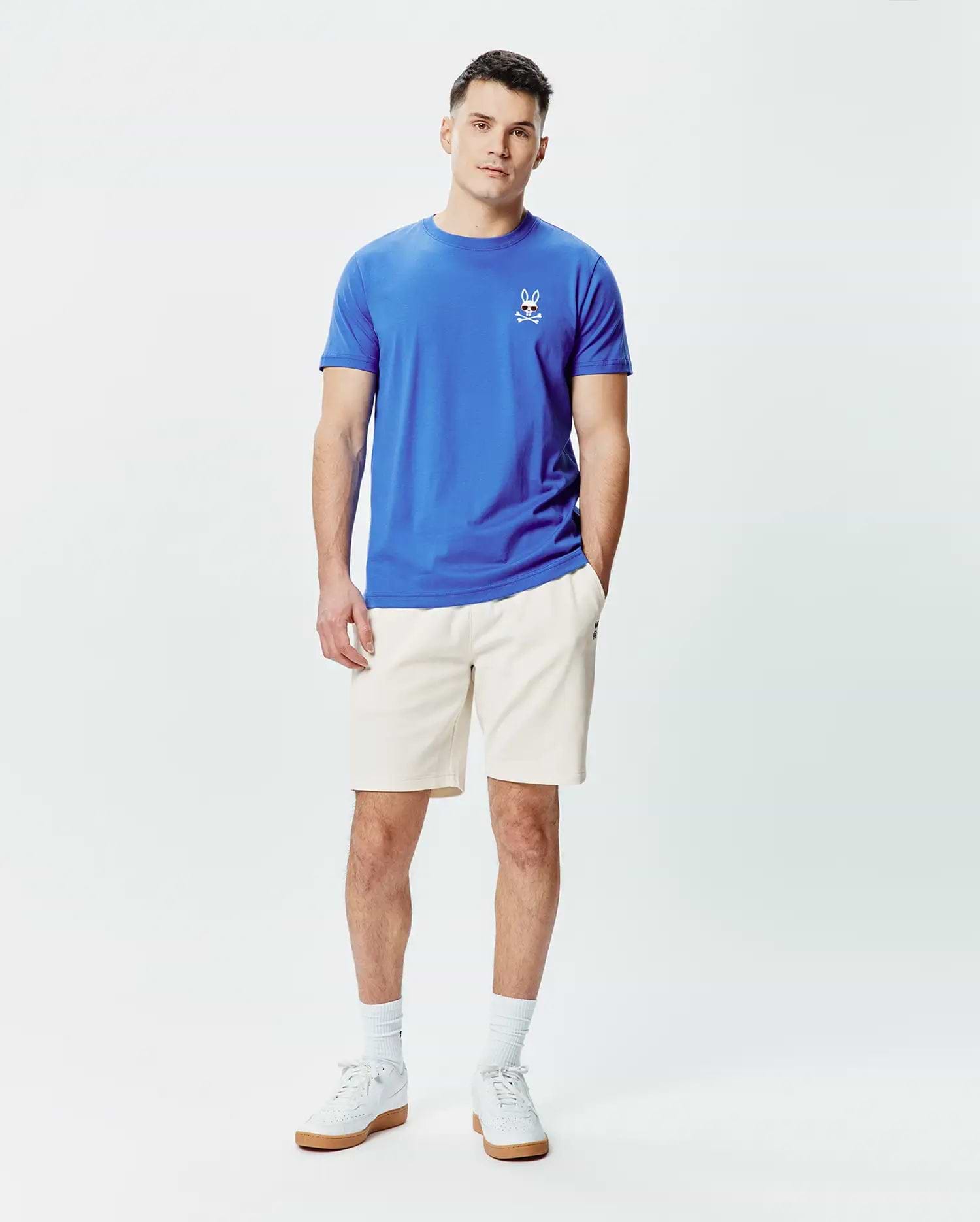 Mens Sunny Chest Graphic Tee and cream shorts. Blue jersey t-shirt with a classic fit, featuring a small embroidered Psycho Bunny skull logo with red eyes and sunglasses on the left chest. Cream relaxed fit shorts.