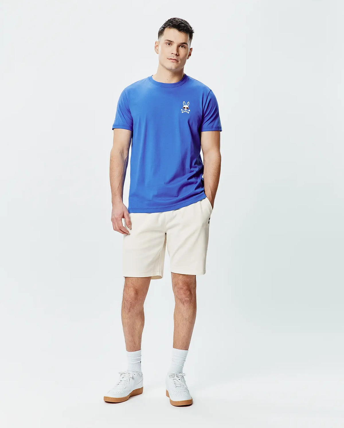Mens Sunny Chest Graphic Tee and cream shorts. Blue jersey t-shirt with a classic fit, featuring a small embroidered Psycho Bunny skull logo with red eyes and sunglasses on the left chest. Cream relaxed fit shorts.