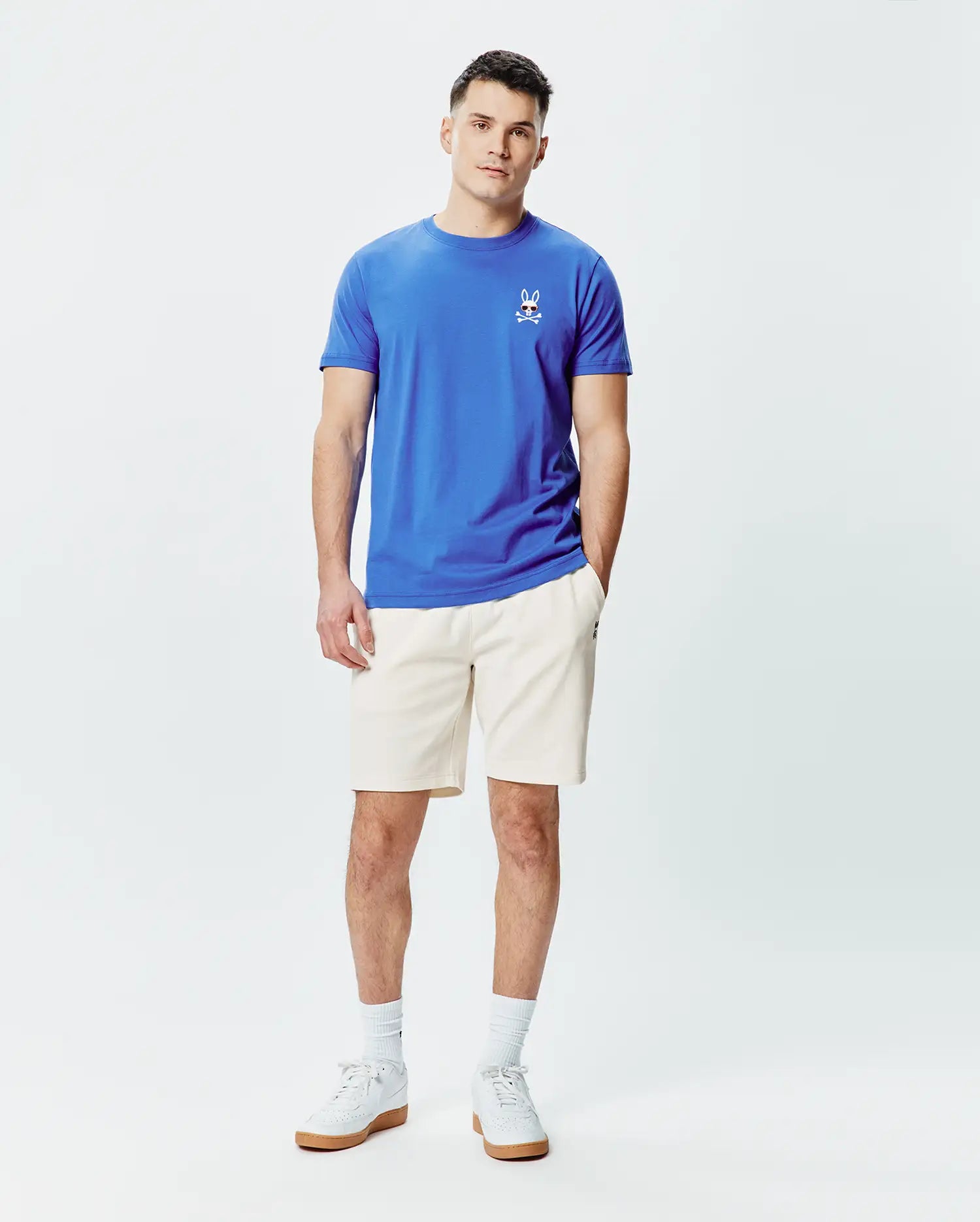 Mens Sunny Chest Graphic Tee and cream shorts. Blue jersey t-shirt with a classic fit, featuring a small embroidered Psycho Bunny skull logo with red eyes and sunglasses on the left chest. Cream relaxed fit shorts.