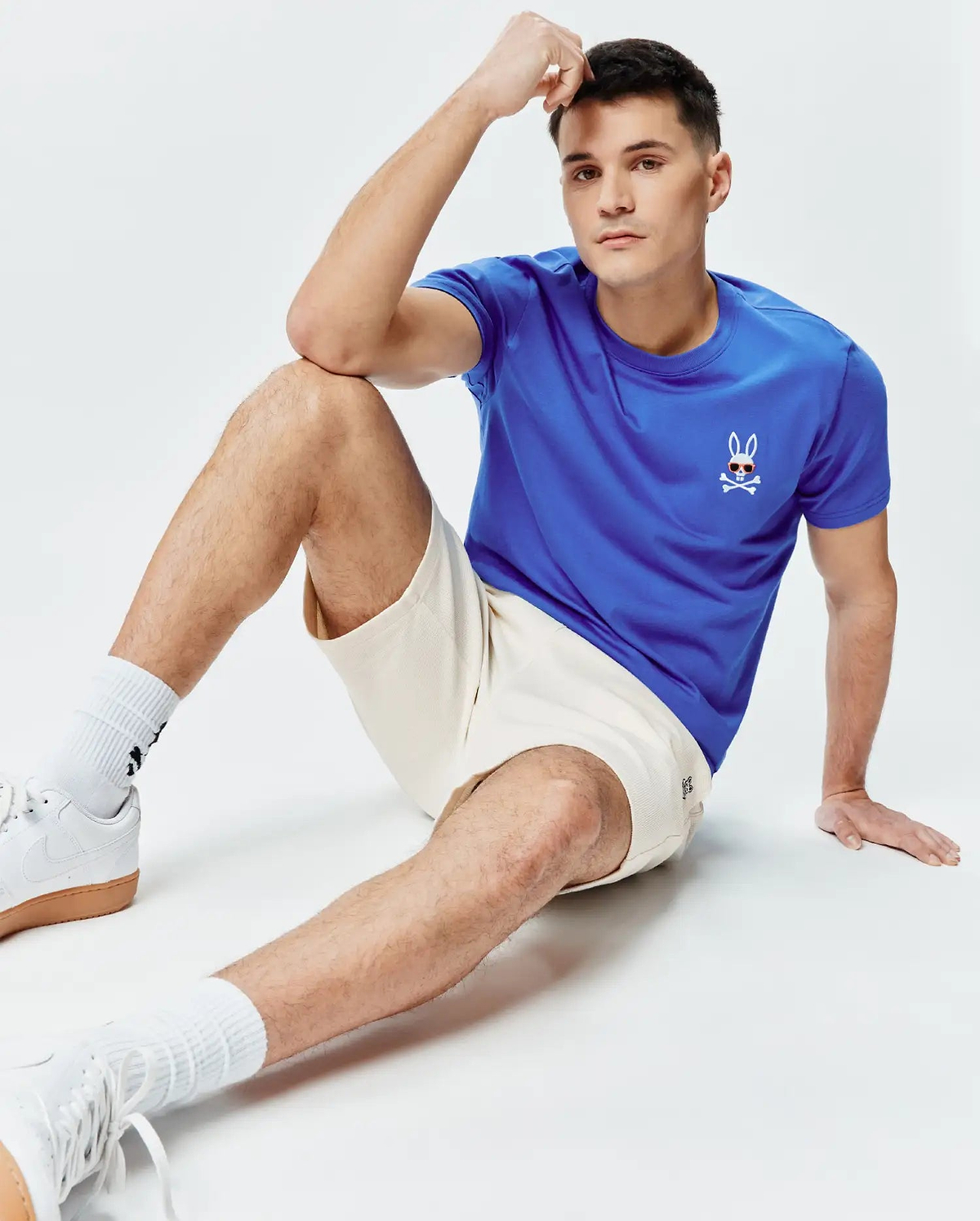 Mens Sunny Chest Graphic Tee in royal blue jersey with a small Psycho Bunny skull logo wearing sunglasses on the left chest, paired with cream textured knit shorts featuring a subtle embroidered bunny logo on the left leg.