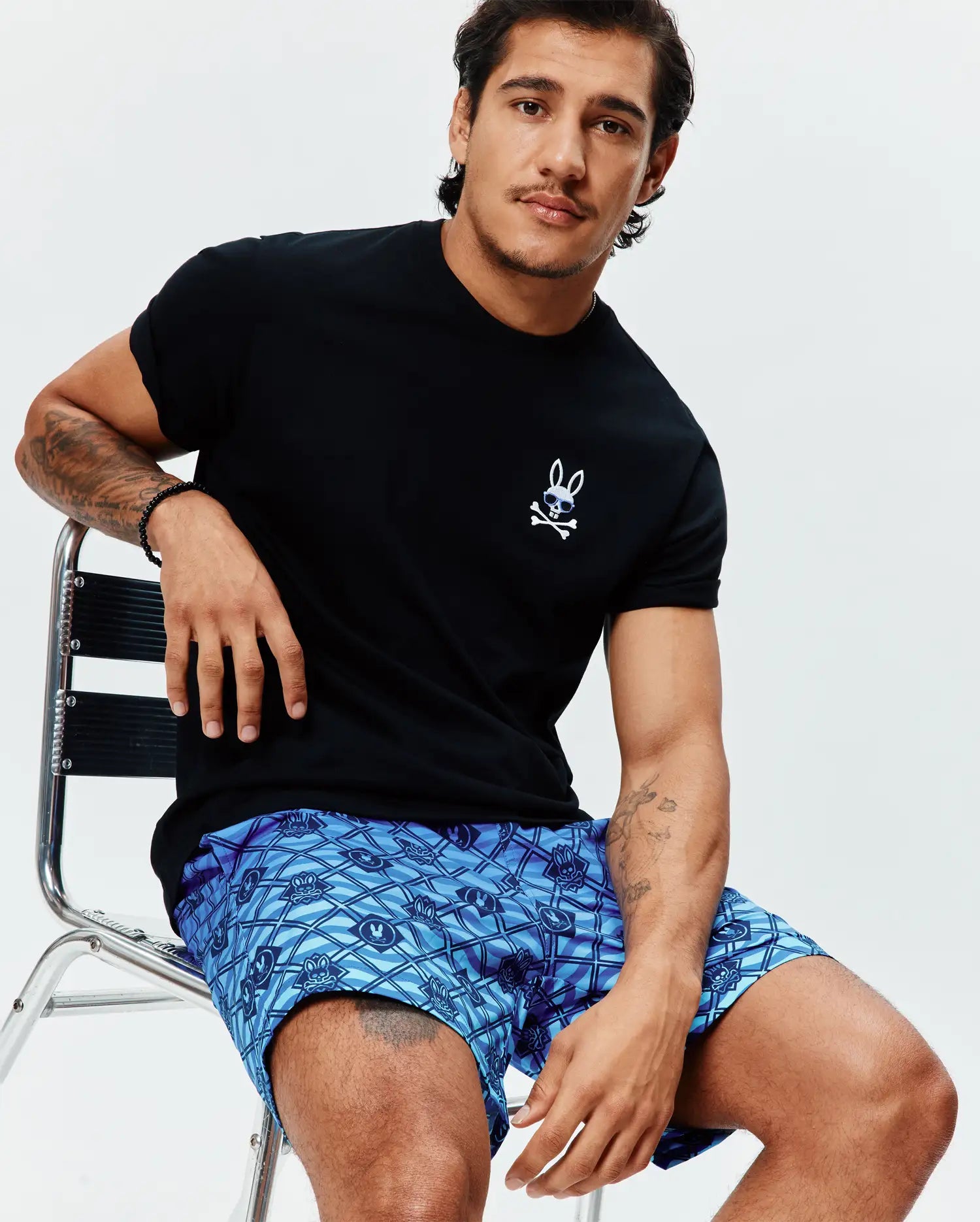 Mens Sunny Chest Graphic Tee, a black jersey t-shirt with a small embroidered white Psycho Bunny skull with sunglasses, paired with blue patterned shorts featuring repeated bunny skull graphics.
