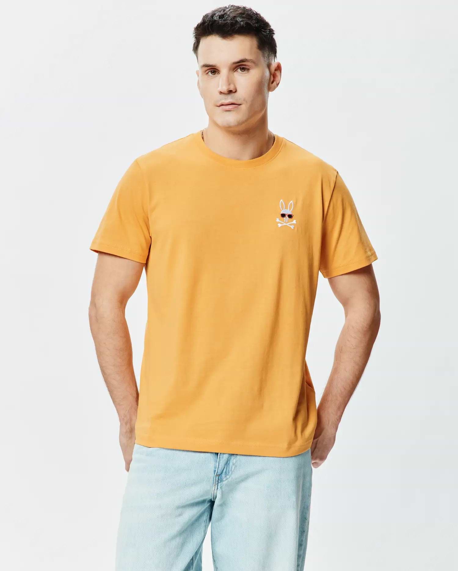 Mens Sunny Chest Graphic Tee. Classic fit mustard yellow smooth jersey t-shirt with a small embroidered Psycho Bunny skull logo wearing red sunglasses and white crossbones on the left chest.