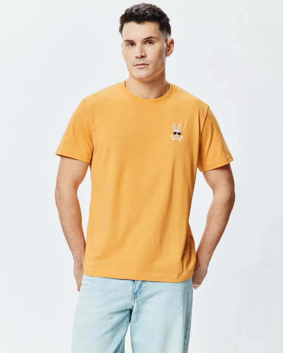 Mens Sunny Chest Graphic Tee. Classic fit mustard yellow smooth jersey t-shirt with a small embroidered Psycho Bunny skull logo wearing red sunglasses and white crossbones on the left chest.