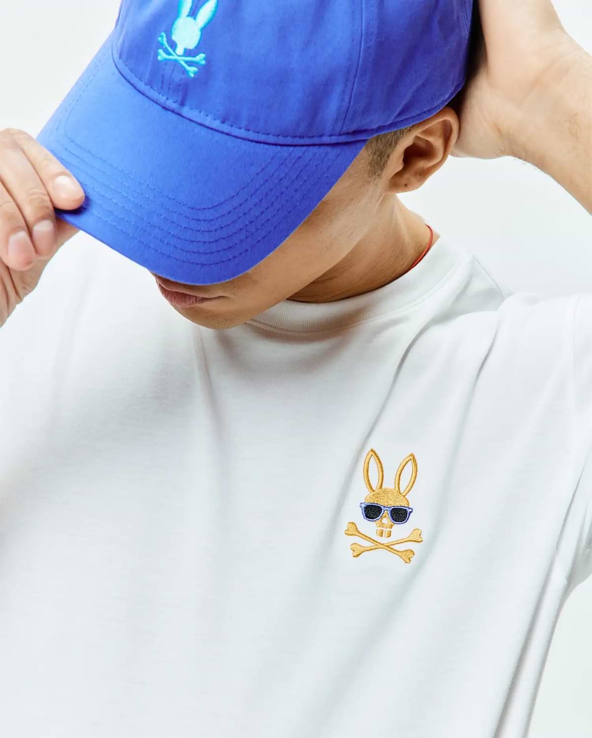 Mens Sunny Chest Graphic Tee, a classic fit white jersey t-shirt featuring a small embroidered gold Psycho Bunny skull with blue sunglasses and crossbones on the left chest, paired with a royal blue cotton twill baseball cap with a light blue embroidered Psycho Bunny logo.