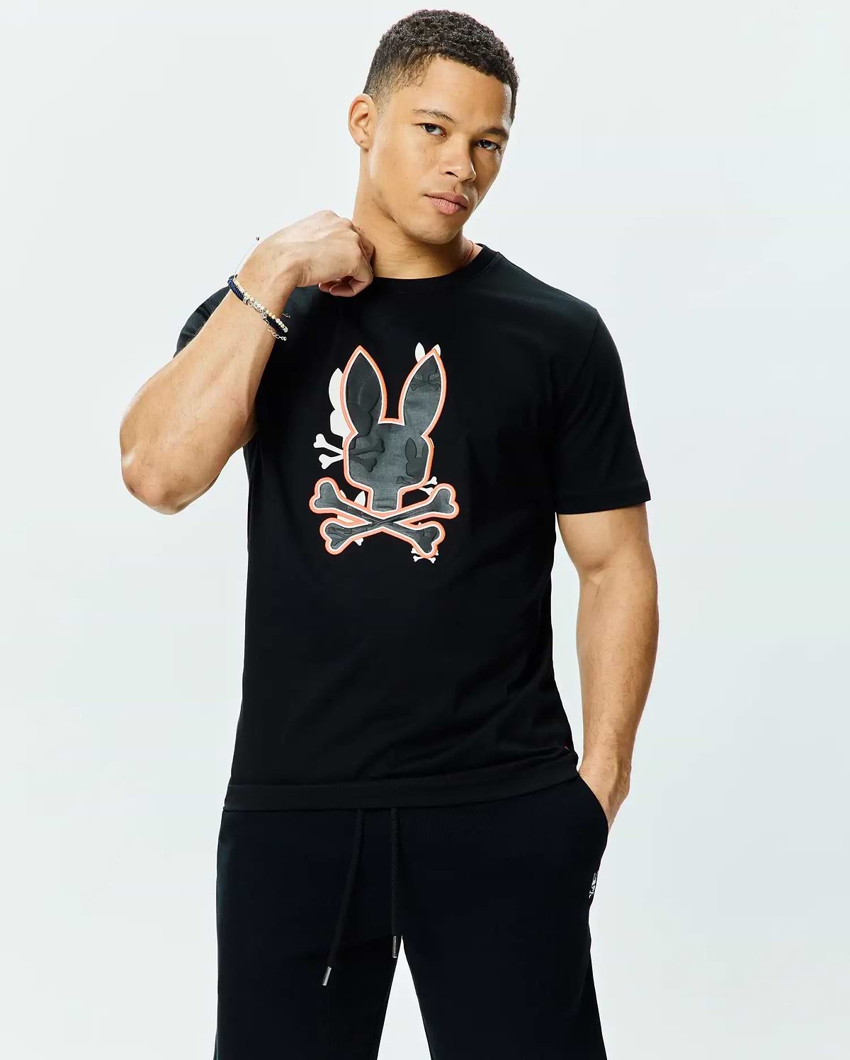 Mens Tanner Bunny Graphic Tee, a black jersey t-shirt with a classic fit, featuring a large central Psycho Bunny skull and crossbones graphic in dark grey camo texture with white and orange outlines.
