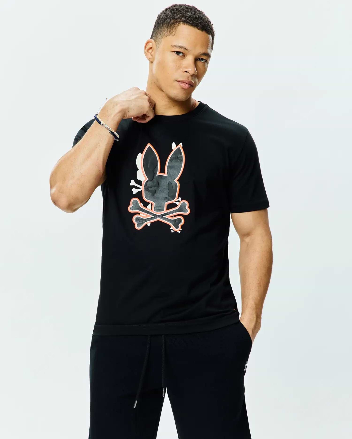 Mens Tanner Bunny Graphic Tee, a black jersey t-shirt with a classic fit, featuring a large central Psycho Bunny skull and crossbones graphic in dark grey camo texture with white and orange outlines.