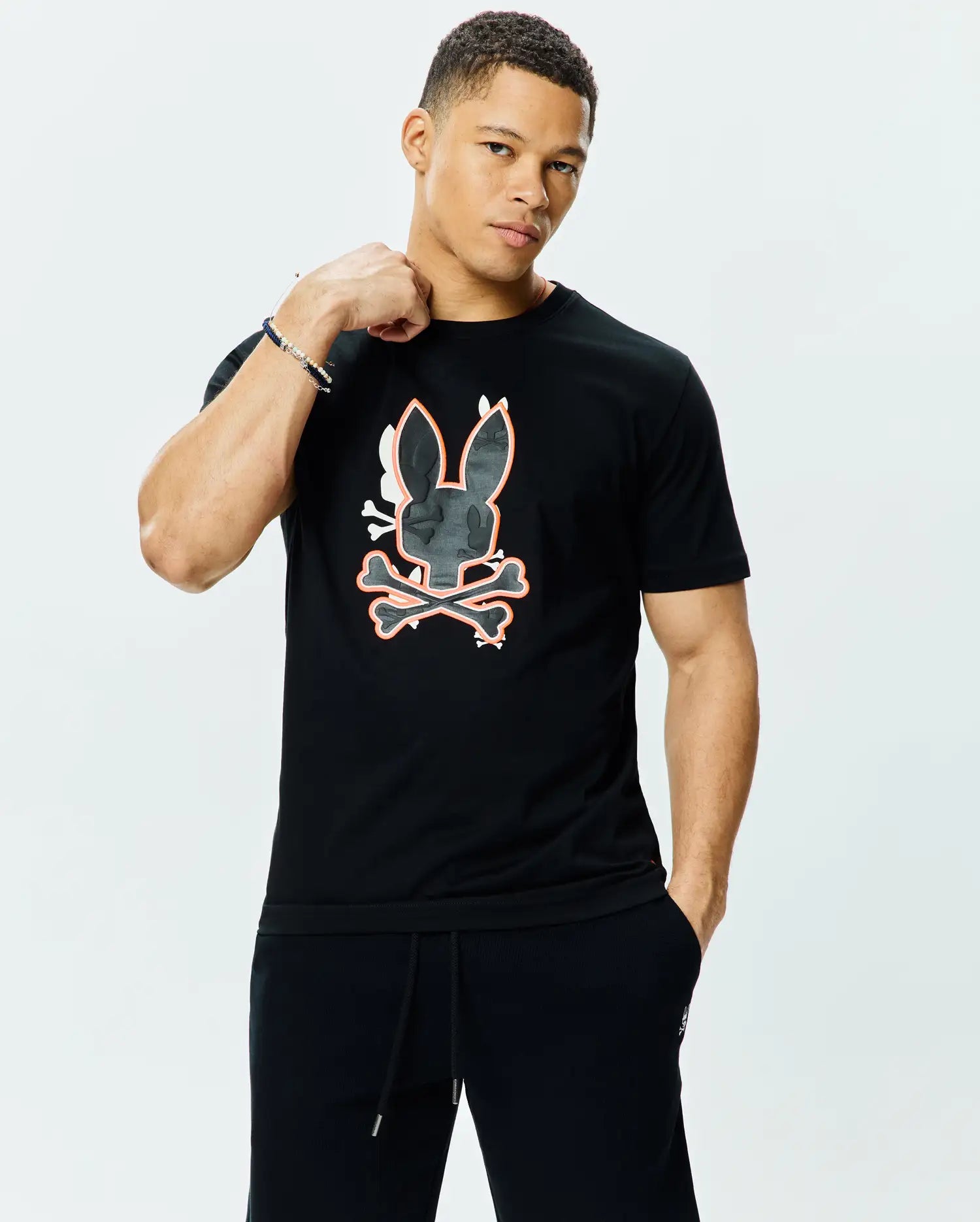 Mens Tanner Bunny Graphic Tee, a black jersey t-shirt with a classic fit, featuring a large central Psycho Bunny skull and crossbones graphic in dark grey camo texture with white and orange outlines.