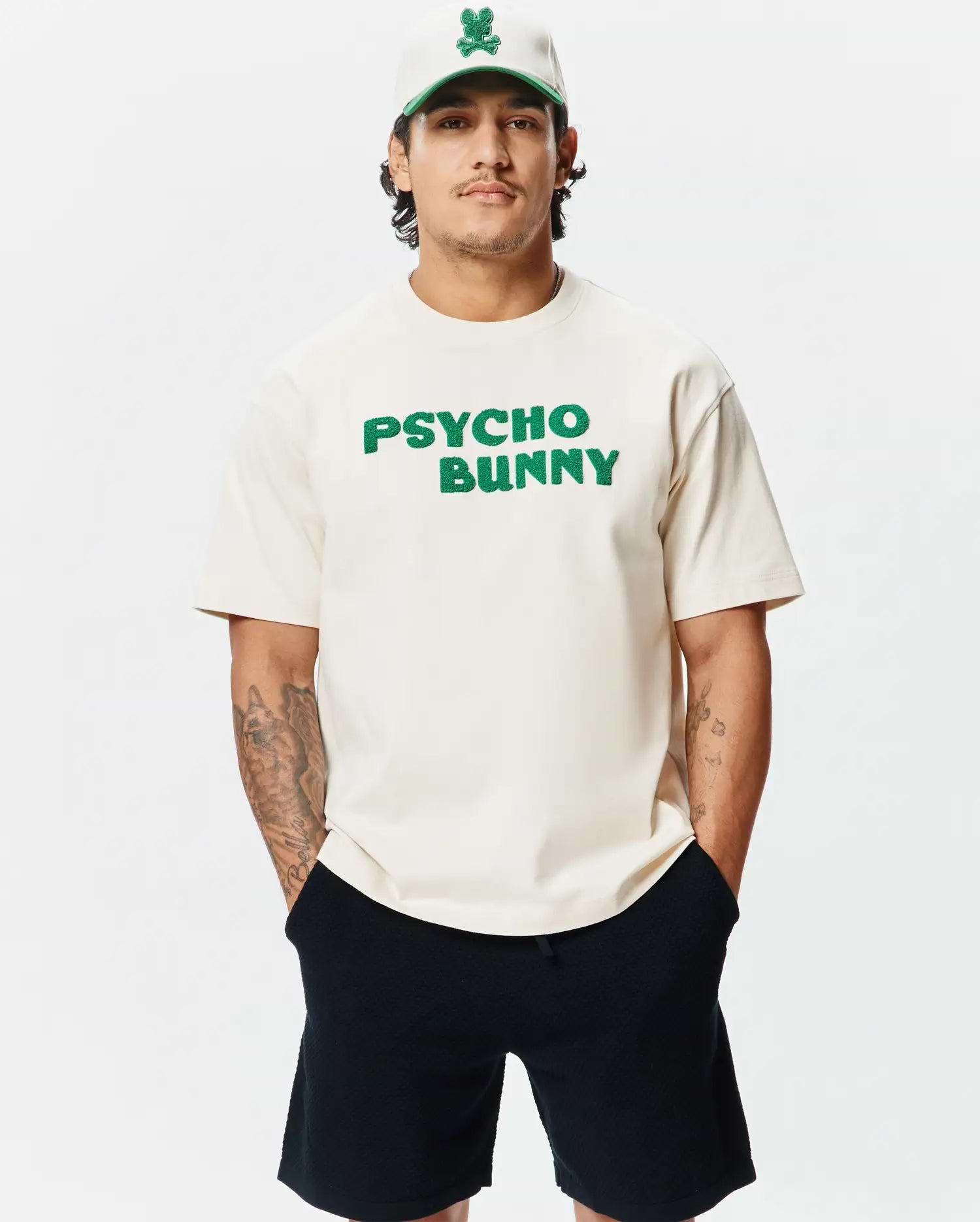 Mens Craig Wordmark Tee in cream jersey with a relaxed fit, featuring a large green textured PSYCHO BUNNY chest logo, complemented by a cream cap with a green brim and embroidered bunny skull.