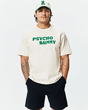 Mens Craig Wordmark Tee in cream jersey with a relaxed fit, featuring a large green textured PSYCHO BUNNY chest logo, complemented by a cream cap with a green brim and embroidered bunny skull.