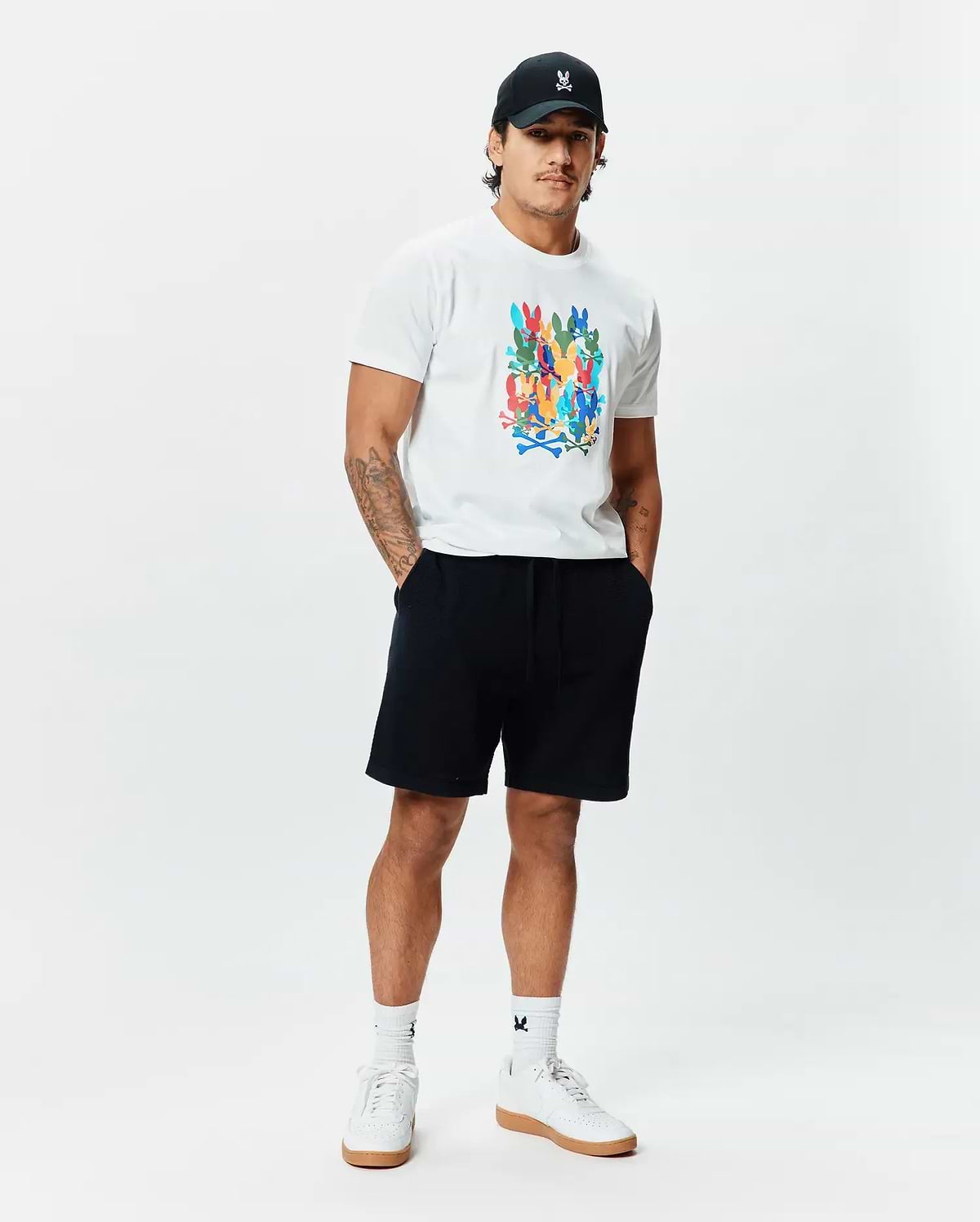 White Mens Tanner Graphic Tee, classic fit, soft jersey, featuring a large multi-color Psycho Bunny skull logo print. Complemented by black French terry shorts, a black cap with an embroidered white bunny logo, and white logo socks.