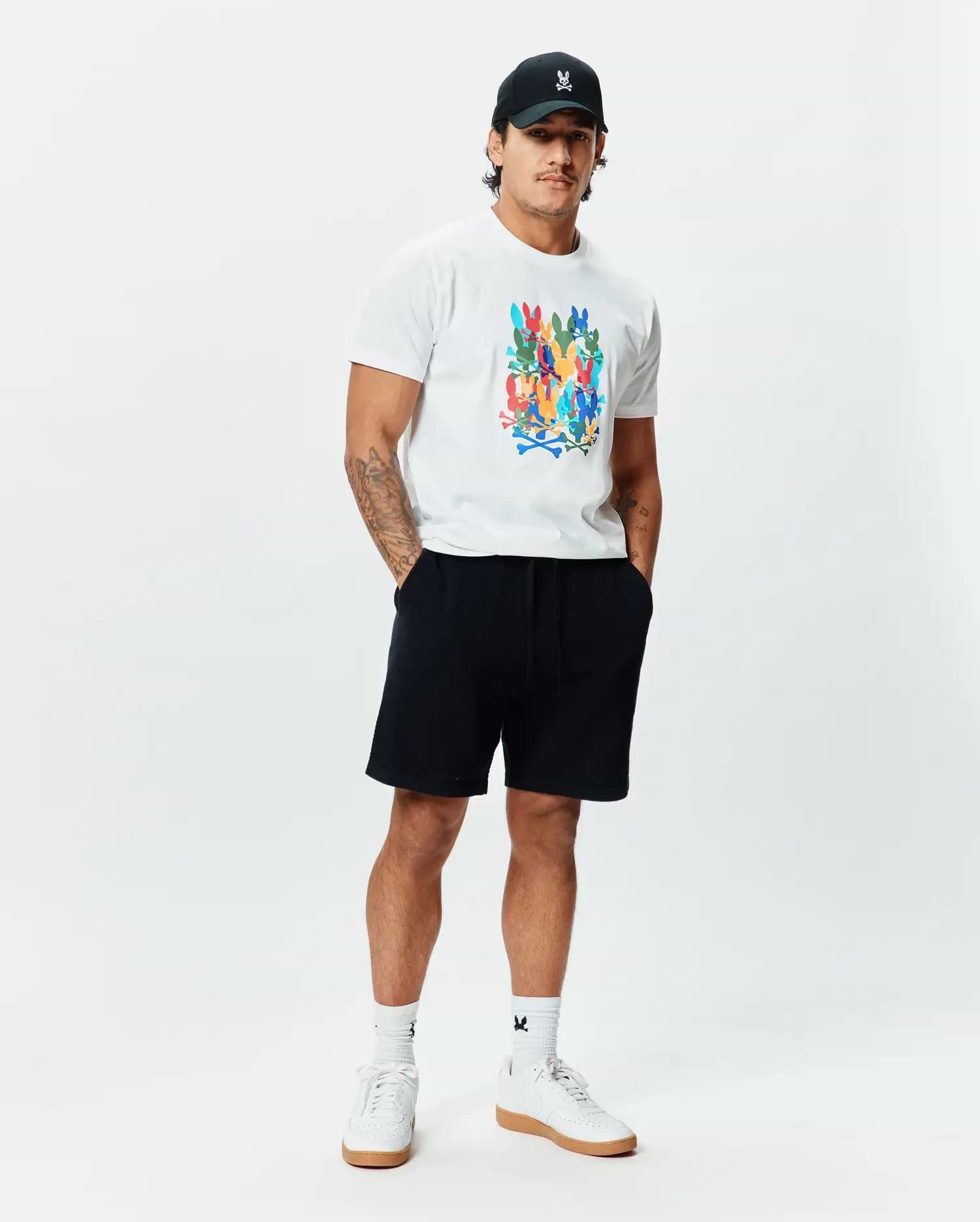 White Mens Tanner Graphic Tee, classic fit, soft jersey, featuring a large multi-color Psycho Bunny skull logo print. Complemented by black French terry shorts, a black cap with an embroidered white bunny logo, and white logo socks.