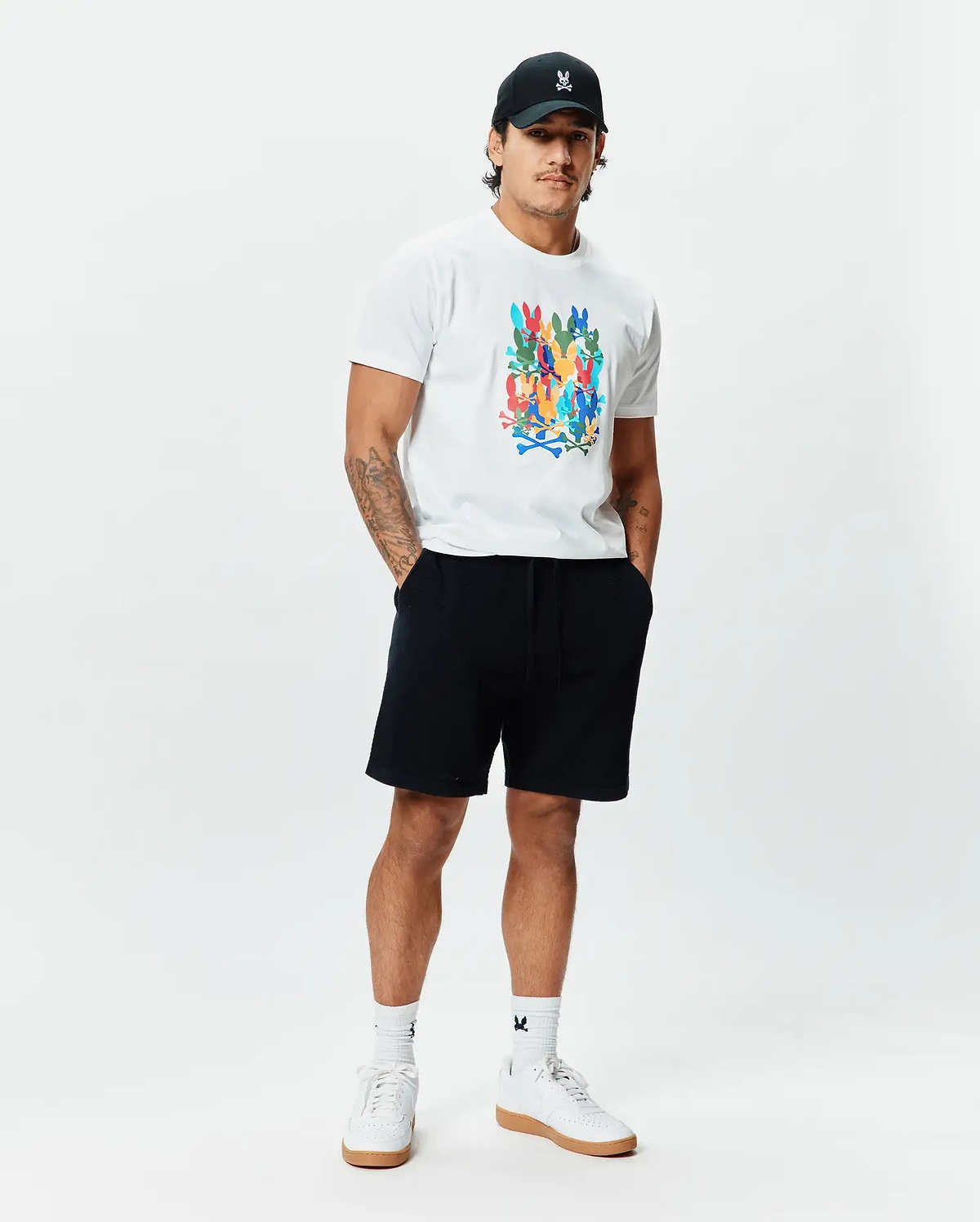 White Mens Tanner Graphic Tee, classic fit, soft jersey, featuring a large multi-color Psycho Bunny skull logo print. Complemented by black French terry shorts, a black cap with an embroidered white bunny logo, and white logo socks.