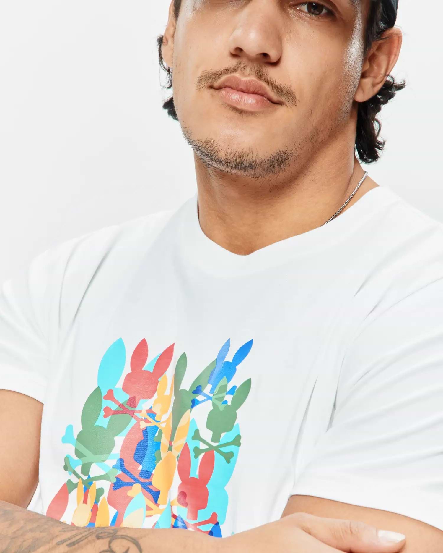 Mens Tanner Graphic Tee, a white classic fit jersey t-shirt with a large, vibrant multi-color chest graphic of overlapping Psycho Bunny skull logos and crossbones.