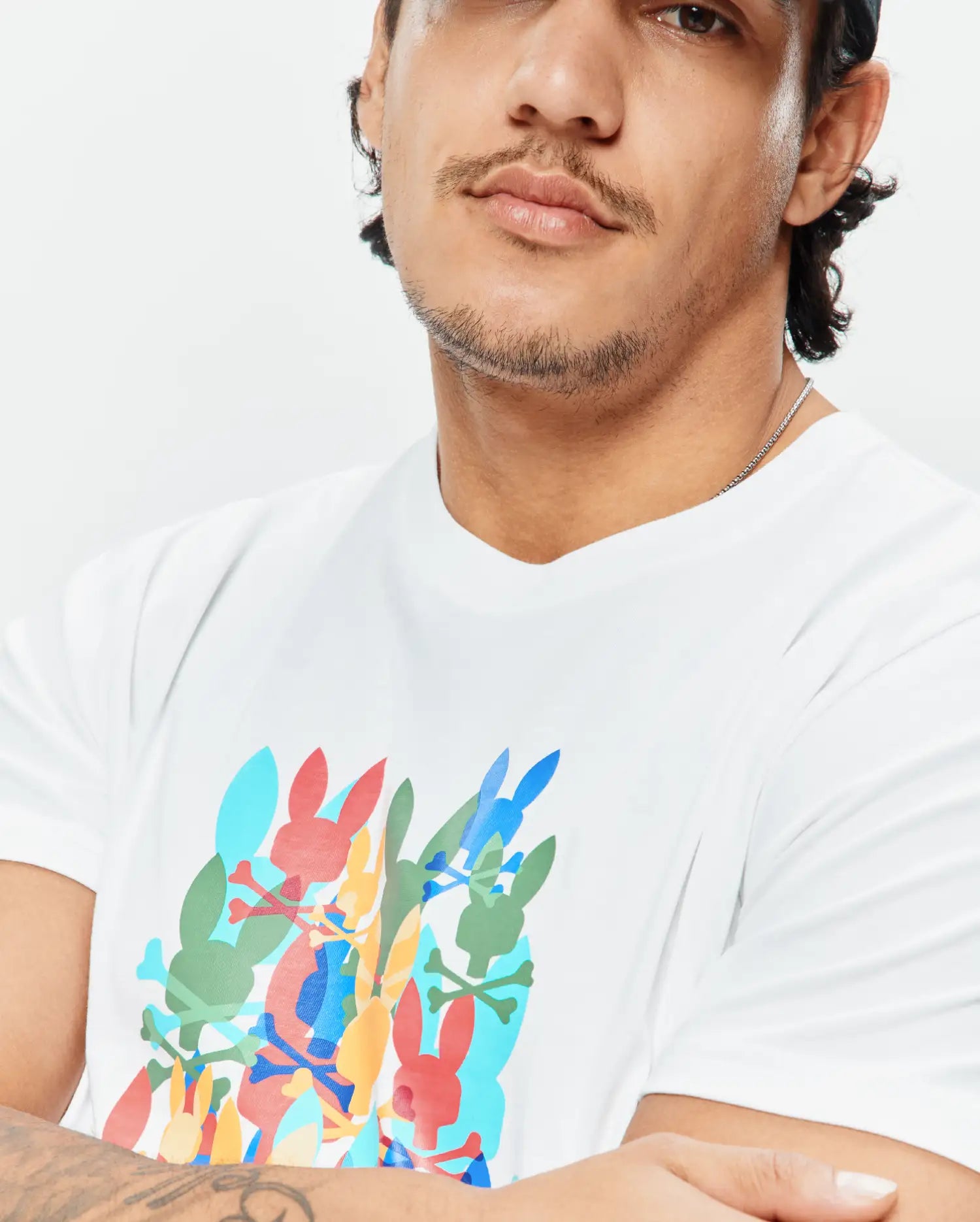 Mens Tanner Graphic Tee, a white classic fit jersey t-shirt with a large, vibrant multi-color chest graphic of overlapping Psycho Bunny skull logos and crossbones.