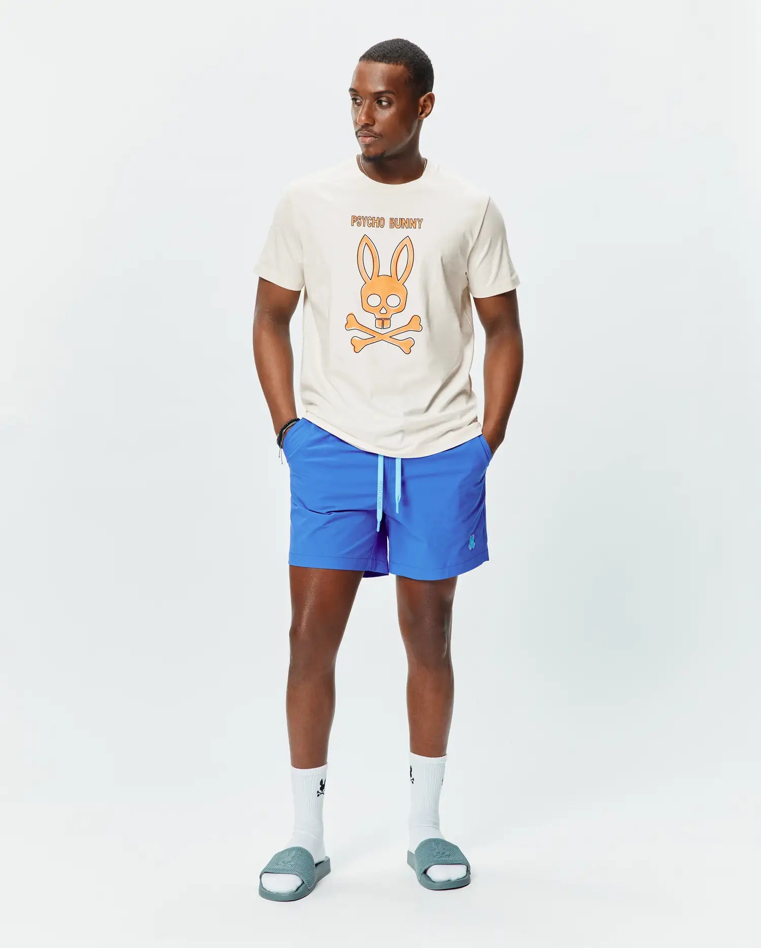 Mens Adler Graphic Tee, a classic fit cream jersey t-shirt with a large orange bunny skull and crossbones graphic on the chest. Paired with bright blue relaxed fit shorts featuring a light blue drawstring and small embroidered bunny logo.