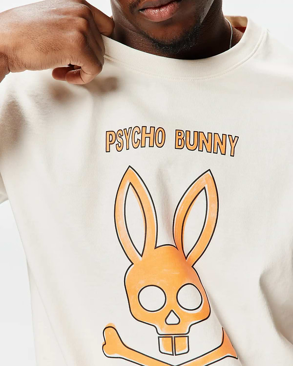 Mens Adler Graphic Tee, a cream classic fit jersey t-shirt featuring a large orange and black Psycho Bunny skull graphic and PSYCHO BUNNY text on the chest.