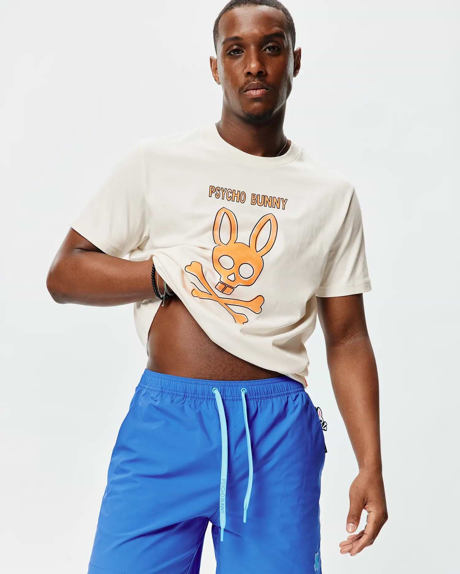 Mens Adler Graphic Tee in cream jersey with a large orange Psycho Bunny skull graphic and text, paired with bright blue relaxed-fit shorts featuring a light blue drawstring and small embroidered bunny logo.
