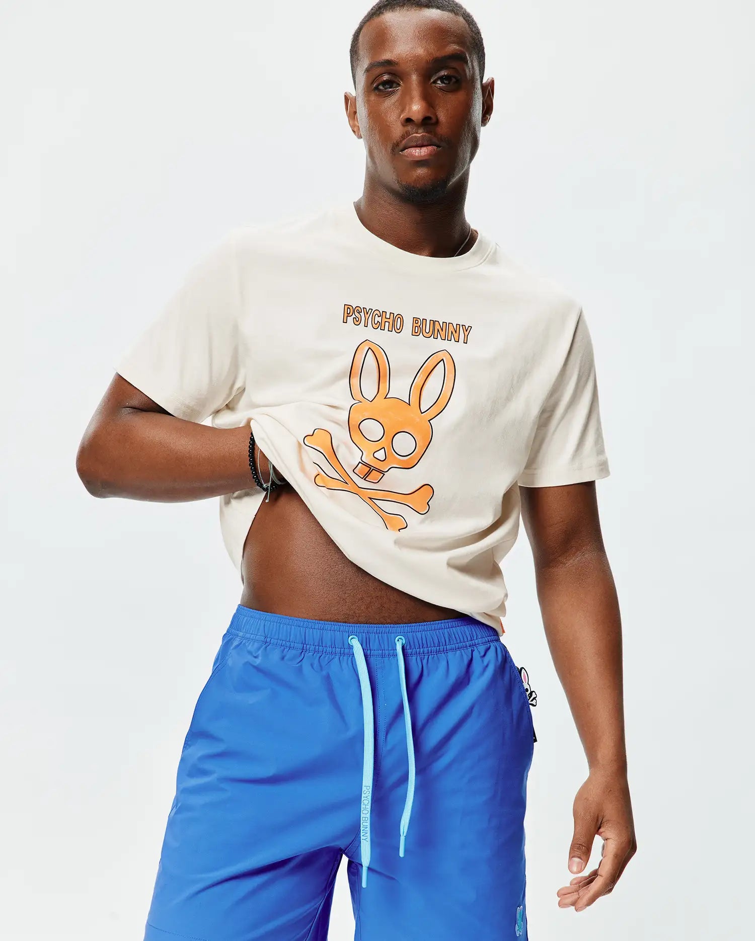 Mens Adler Graphic Tee in cream jersey with a large orange Psycho Bunny skull graphic and text, paired with bright blue relaxed-fit shorts featuring a light blue drawstring and small embroidered bunny logo.