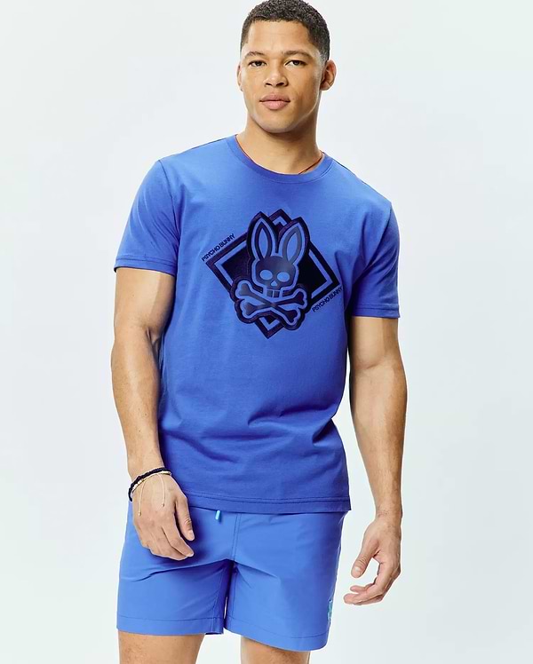 Mens Tobias Graphic Tee, a classic fit royal blue jersey t-shirt with a large dark blue Psycho Bunny skull and crossbones logo graphic in a diamond on the chest, and subtle orange collar trim.