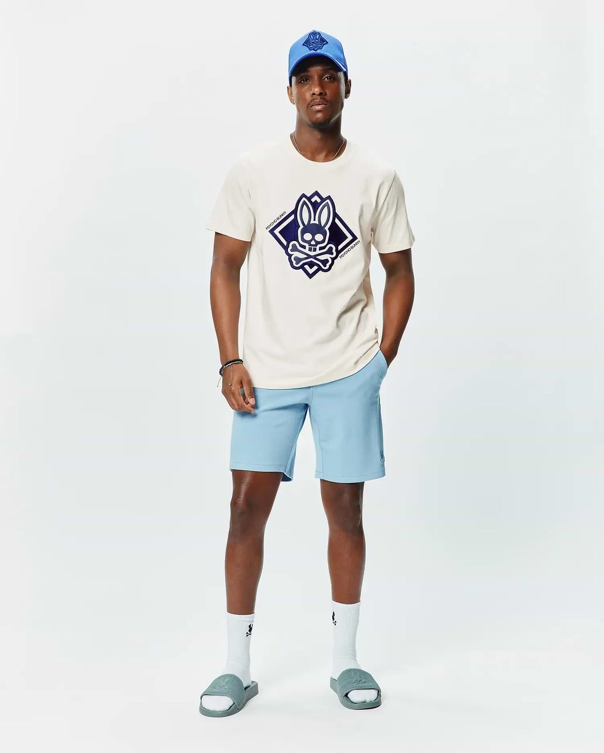 Mens Tobias Graphic Tee in ecru jersey with a large navy diamond Psycho Bunny skull logo on the chest, paired with light blue shorts featuring a small embroidered logo, a royal blue cap, white socks, and grey slides.