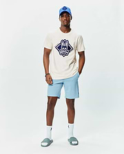 Mens Tobias Graphic Tee in ecru jersey with a large navy diamond Psycho Bunny skull logo on the chest, paired with light blue shorts featuring a small embroidered logo, a royal blue cap, white socks, and grey slides.