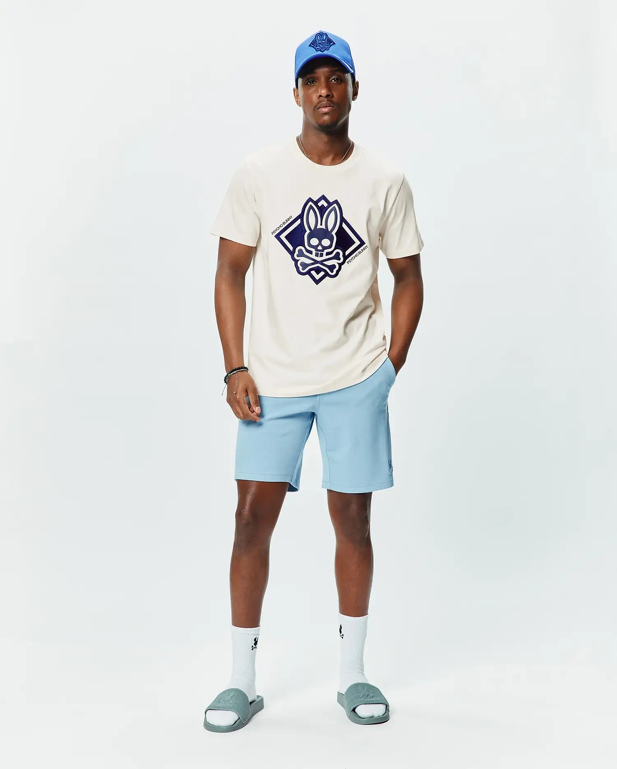 Mens Tobias Graphic Tee in ecru jersey with a large navy diamond Psycho Bunny skull logo on the chest, paired with light blue shorts featuring a small embroidered logo, a royal blue cap, white socks, and grey slides.