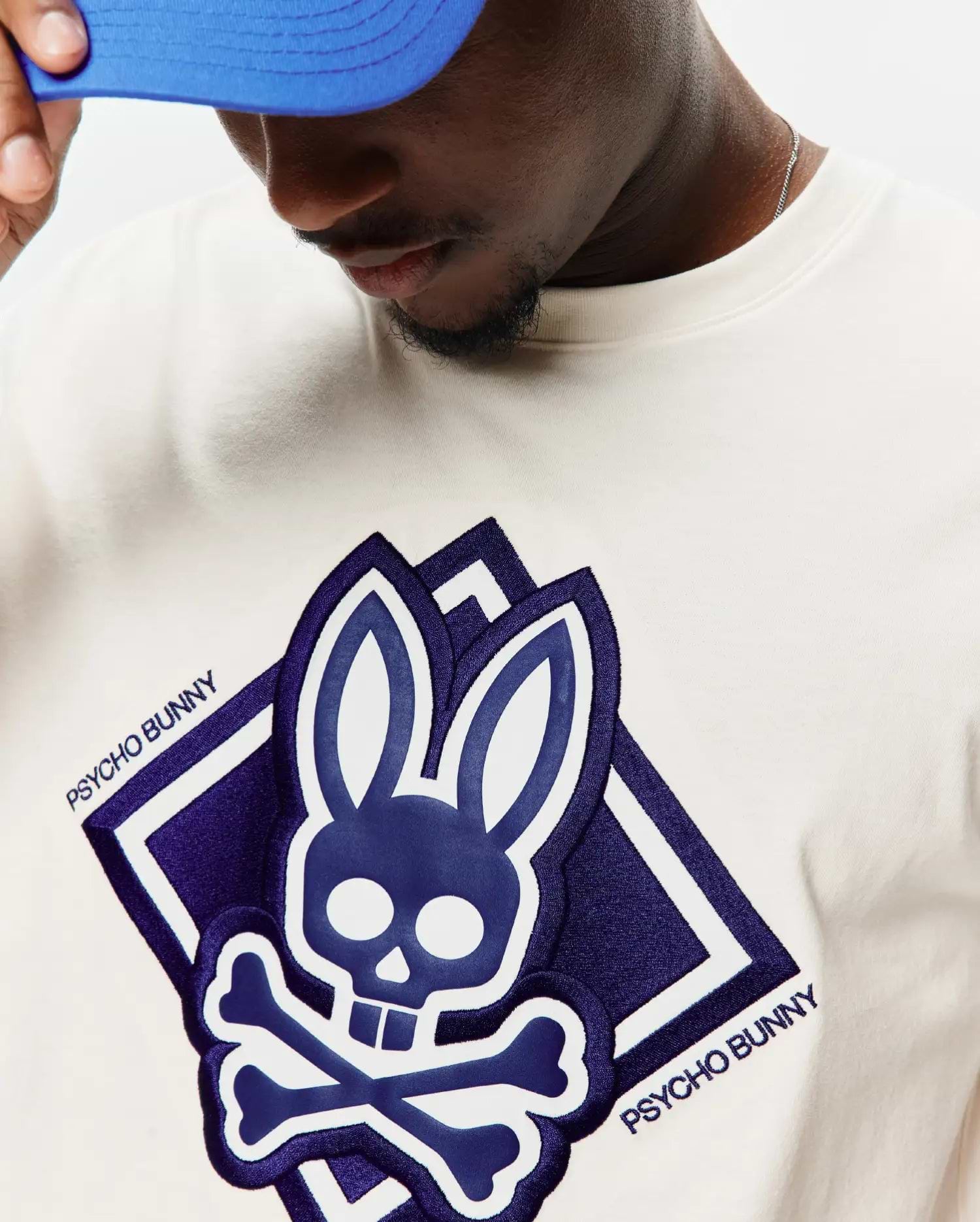 Mens Tobias Graphic Tee, an off-white jersey knit t-shirt with a large central deep purple chenille Psycho Bunny skull and crossbones logo graphic, outlined in white, featuring embroidered PSYCHO BUNNY text.