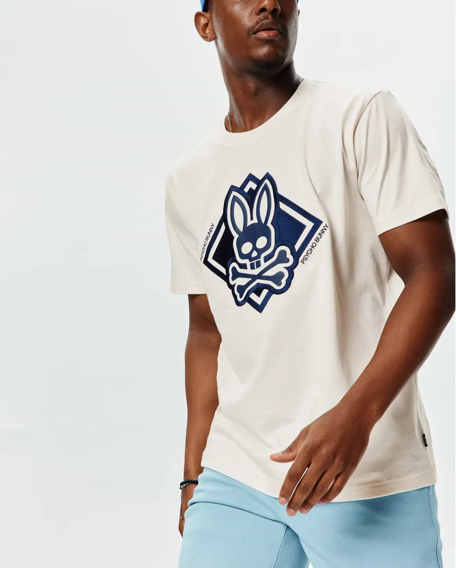 Mens Tobias Graphic Tee, a classic fit ecru jersey t-shirt featuring a large navy blue Psycho Bunny skull and crossbones logo within a diamond graphic on the chest, with Psycho Bunny text vertically on the right side.