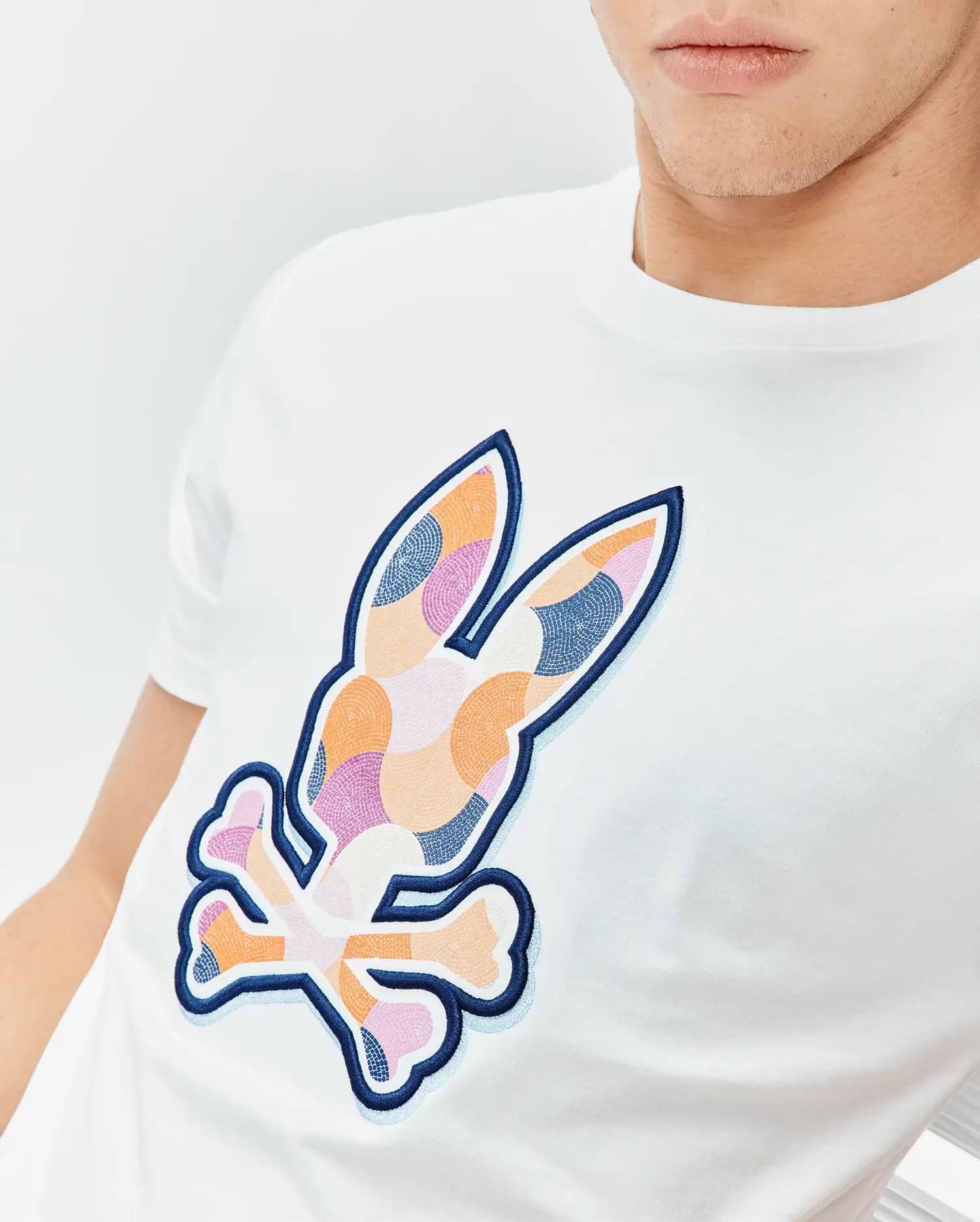 Mens Ethan Graphic Tee: White classic fit jersey t-shirt featuring a large, central embroidered Psycho Bunny skull logo with a colorful mosaic pattern in orange, pink, and blue, outlined in dark blue with a light blue shadow. (3)