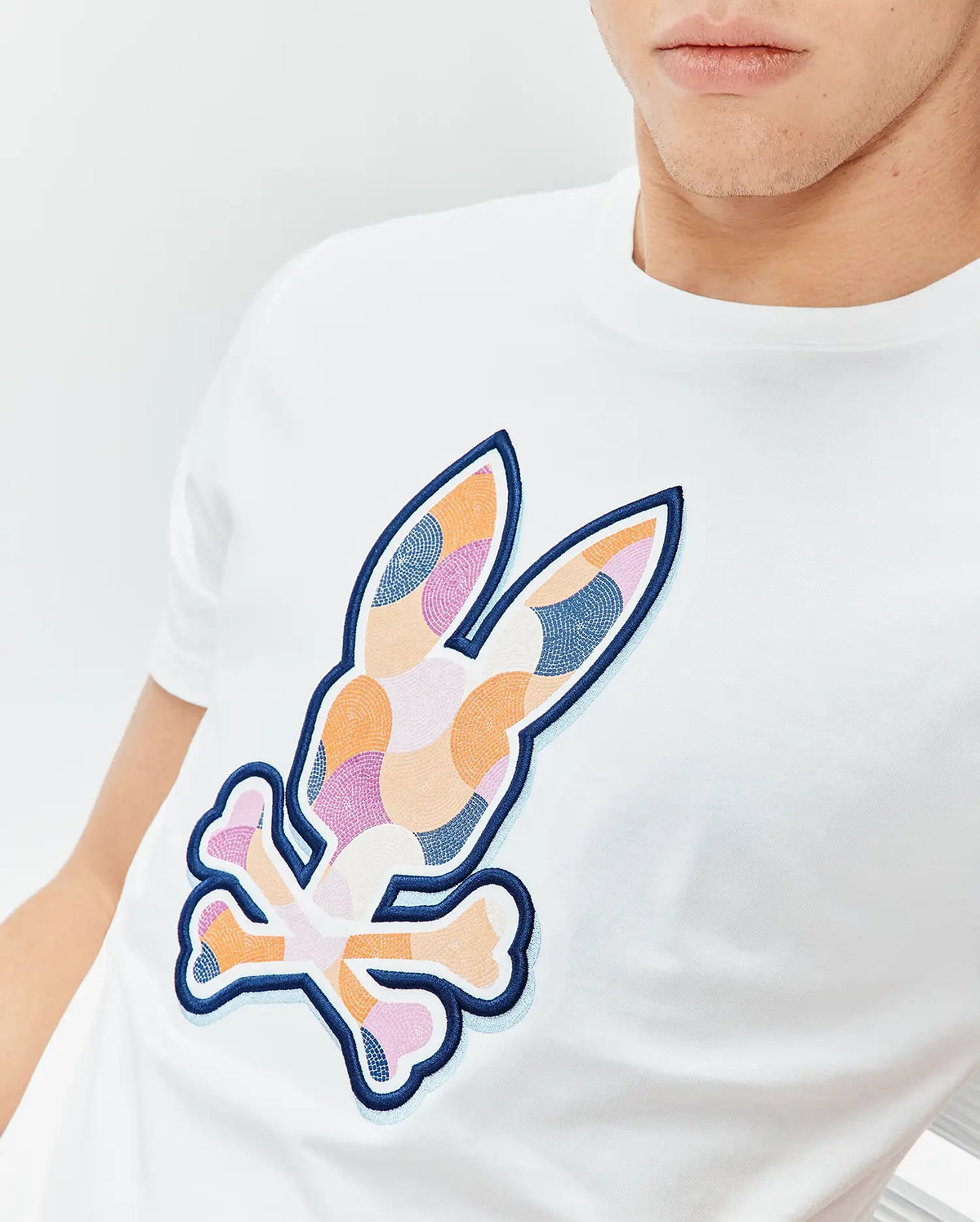Mens Ethan Graphic Tee: White classic fit jersey t-shirt featuring a large, central embroidered Psycho Bunny skull logo with a colorful mosaic pattern in orange, pink, and blue, outlined in dark blue with a light blue shadow. (3)