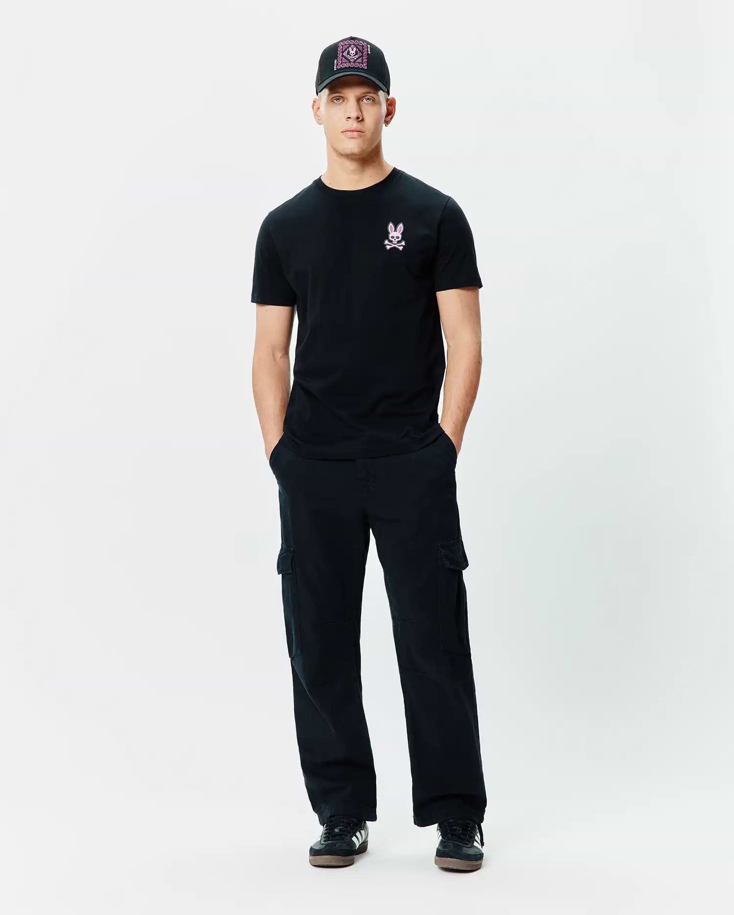 Black Mens Frank Back Graphic Tee, classic fit, with a small pink embroidered Psycho Bunny skull logo on the left chest, complemented by black relaxed cargo pants and a black baseball cap featuring a vibrant pink patterned front.
