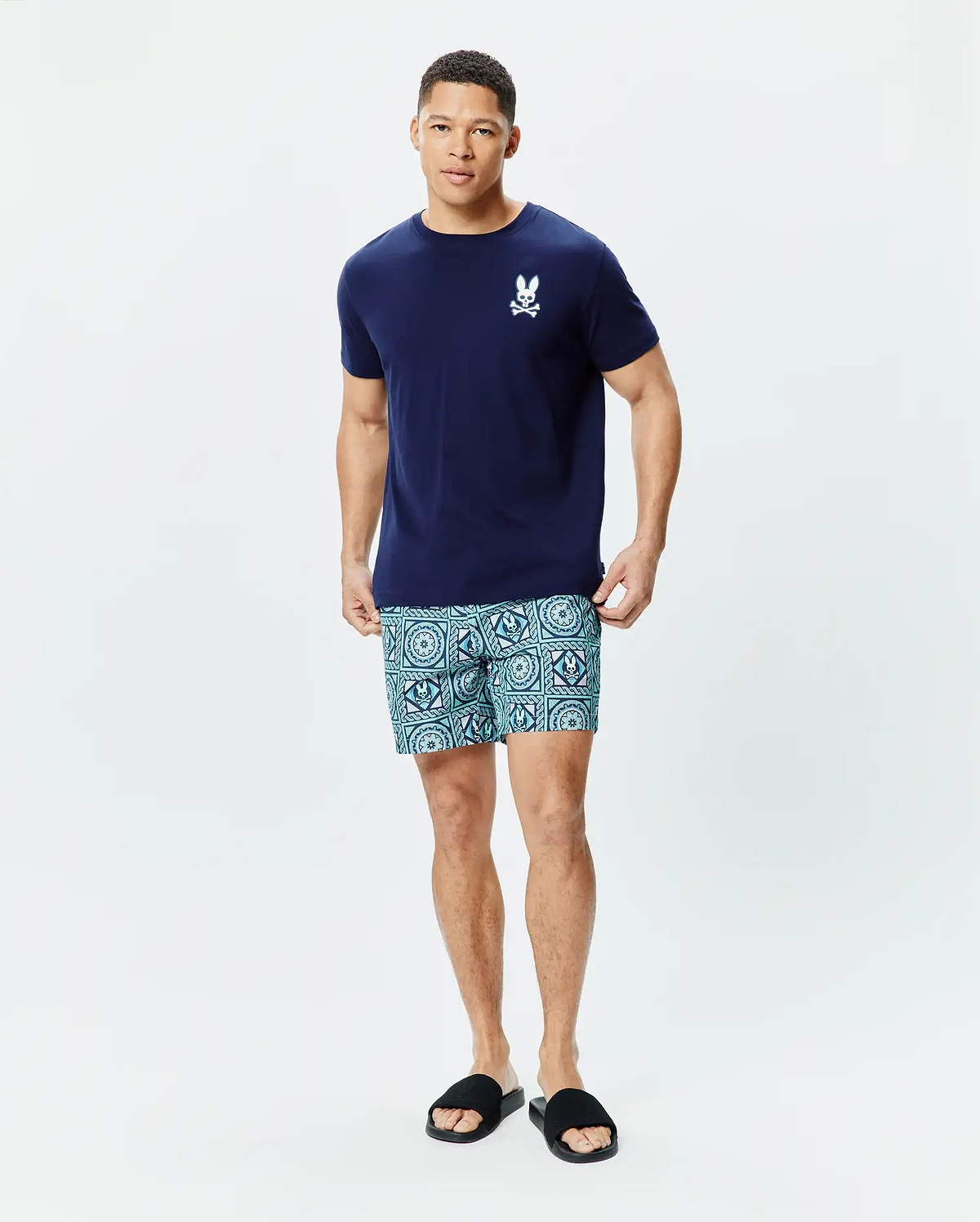 Mens Frank Back Graphic Tee in navy blue jersey, classic fit, with a small white embroidered Psycho Bunny skull logo on the left chest, paired with light blue patterned shorts featuring geometric and subtle skull motifs.