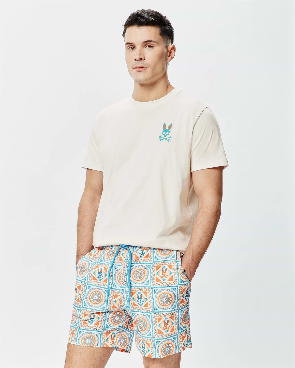 Cream Mens Frank Back Graphic Tee, a classic fit jersey t-shirt with a small embroidered teal and orange Psycho Bunny skull logo on the left chest, paired with patterned swim shorts. (1)