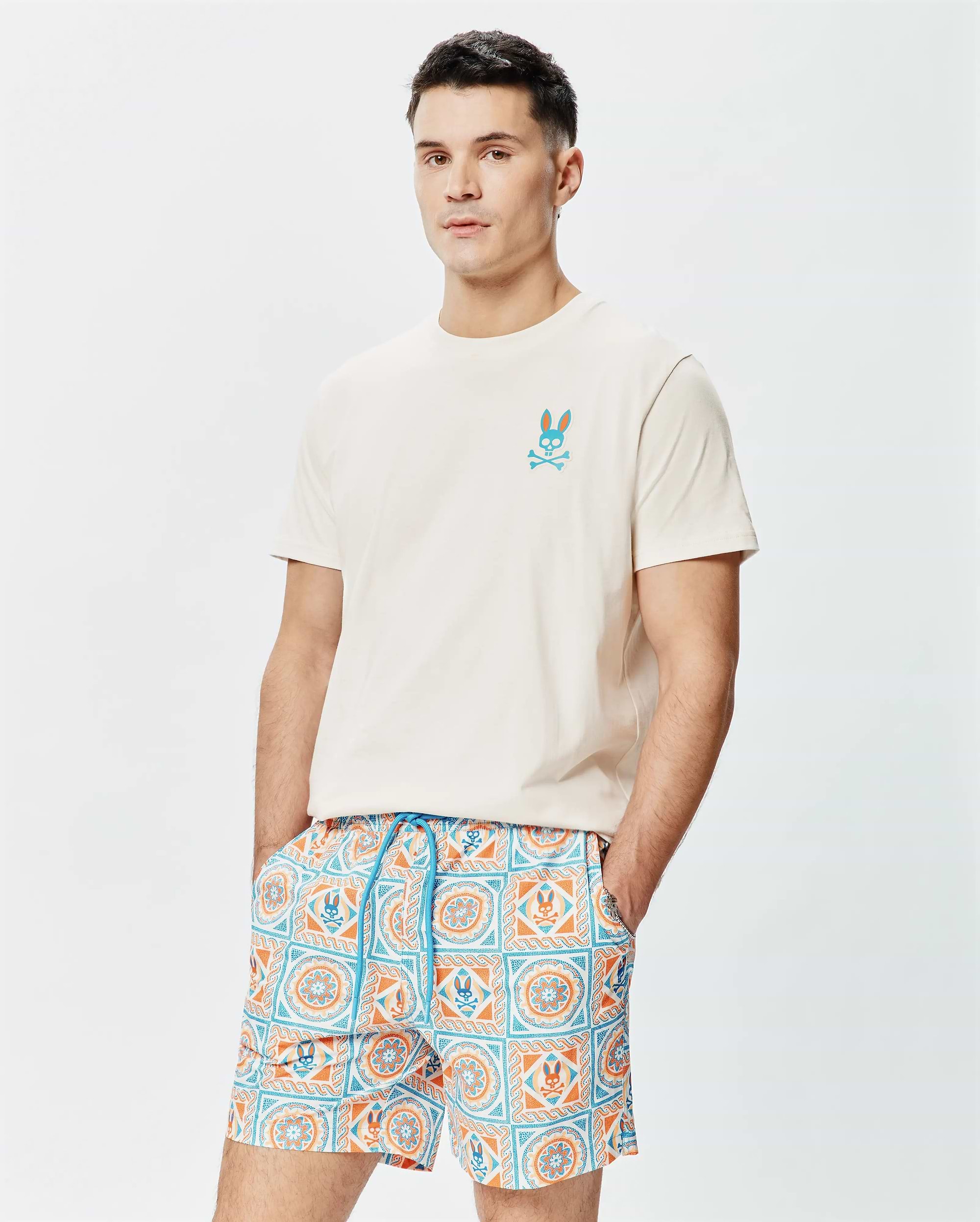 Cream Mens Frank Back Graphic Tee, a classic fit jersey t-shirt with a small embroidered teal and orange Psycho Bunny skull logo on the left chest, paired with patterned swim shorts. (1)